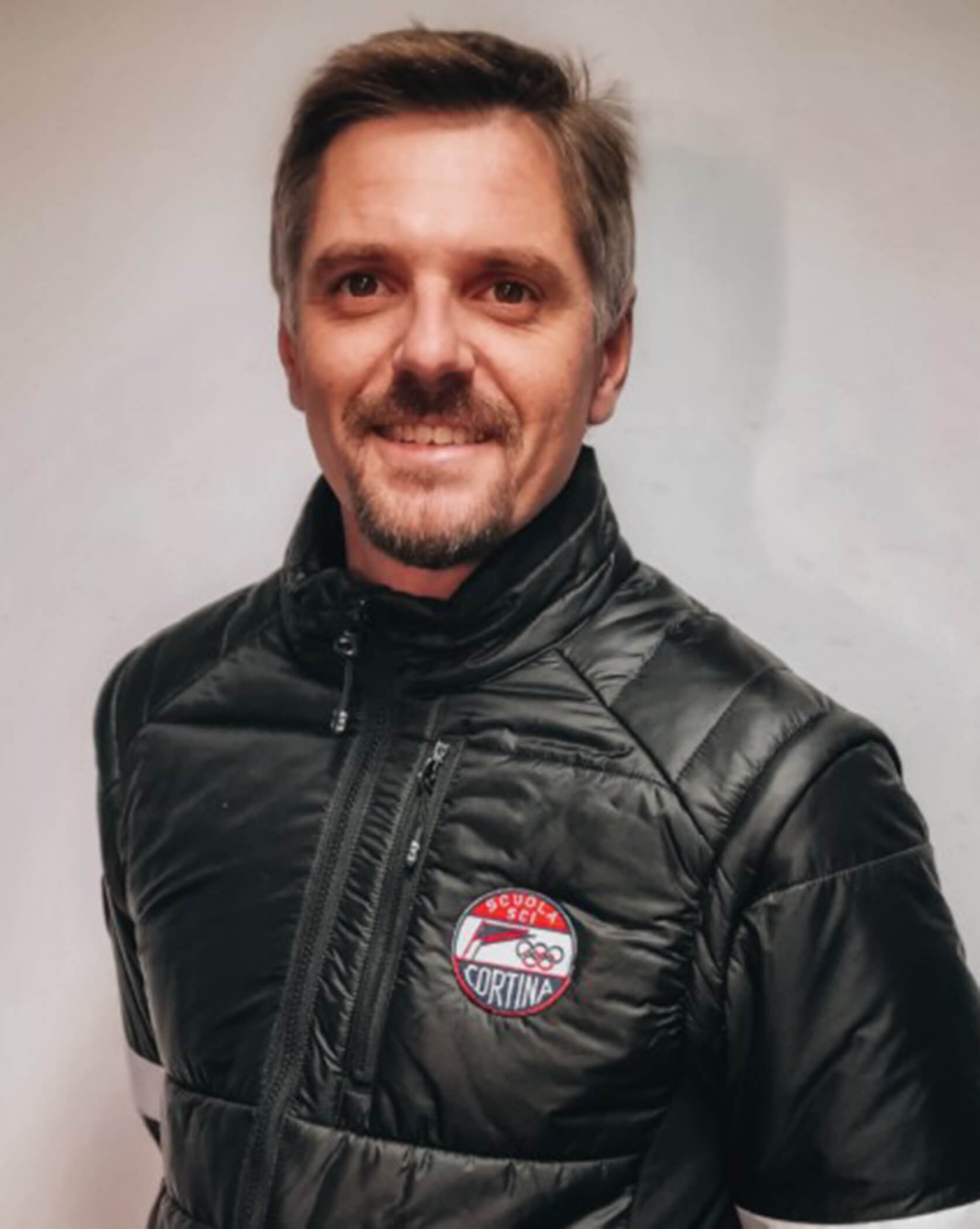 Man in a black puffer jacket with a Cortina badge, standing against a plain white background, smiling. - Scuola Sci Cortina
