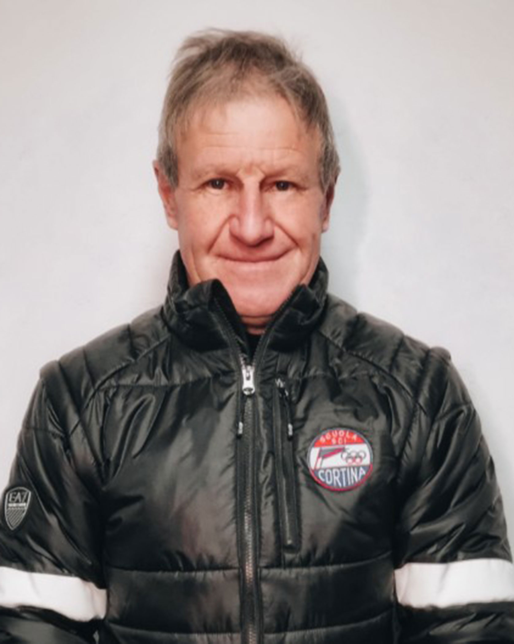 Older man in a black puffy jacket with a Cortina patch, standing against a plain light background. - Scuola Sci Cortina