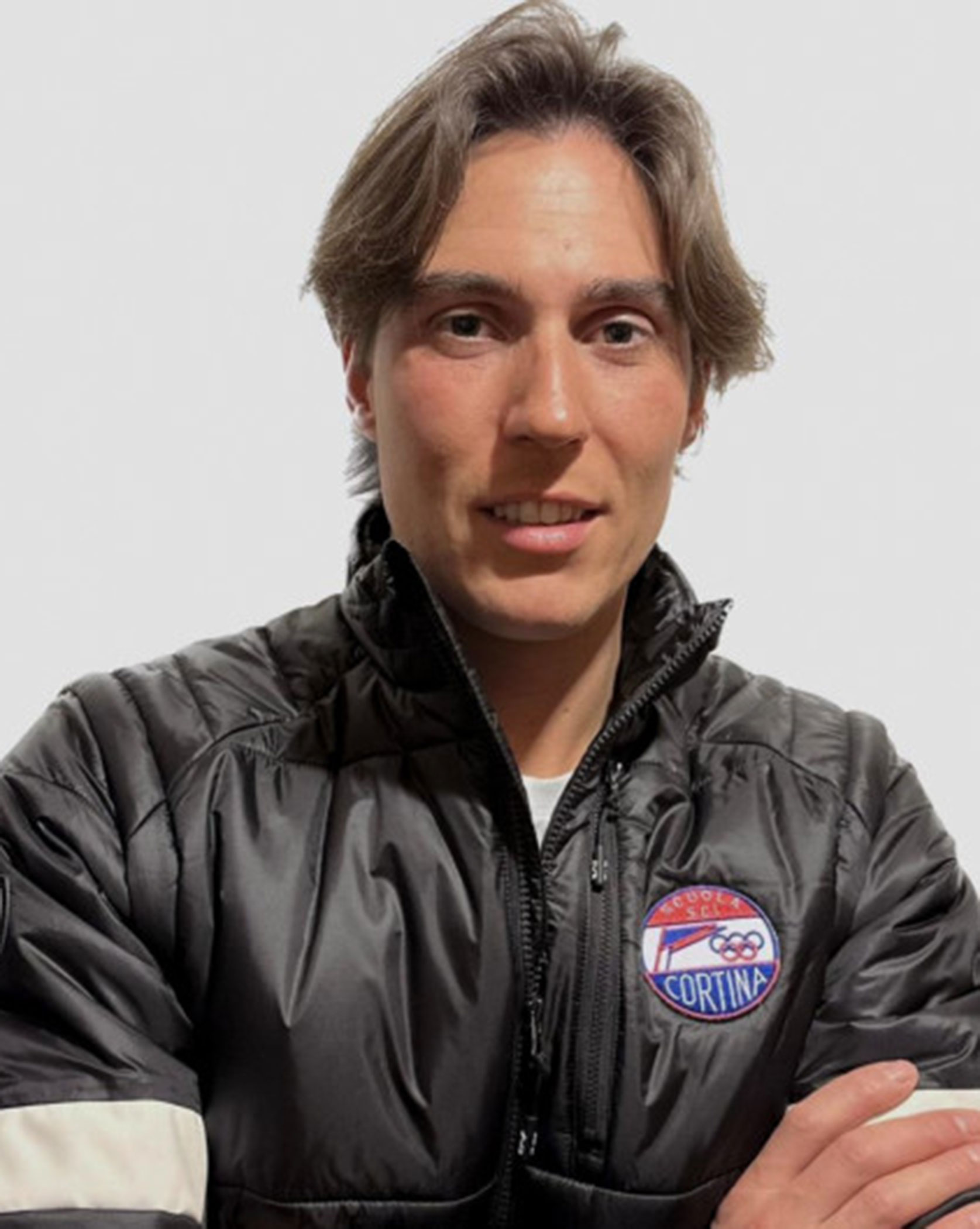 Person in a black jacket with a Cortina patch, standing with arms folded, neutral background. - Scuola Sci Cortina