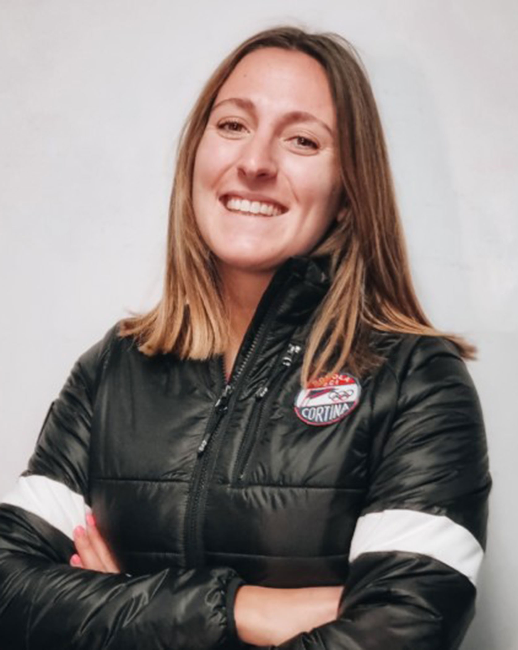 Smiling woman with straight brown hair in a black jacket with a Cortina logo, standing against a plain wall. - Scuola Sci Cortina