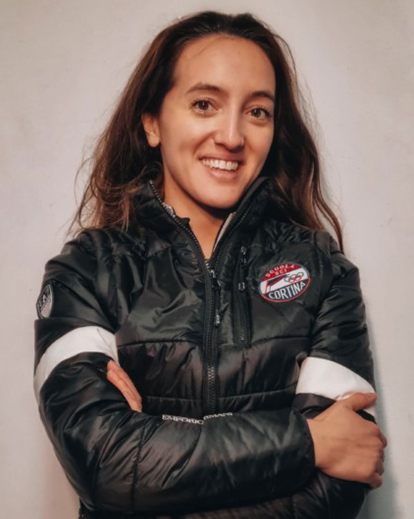 Person in a black puffer jacket with crossed arms, smiling at the camera against a plain background. - Scuola Sci Cortina