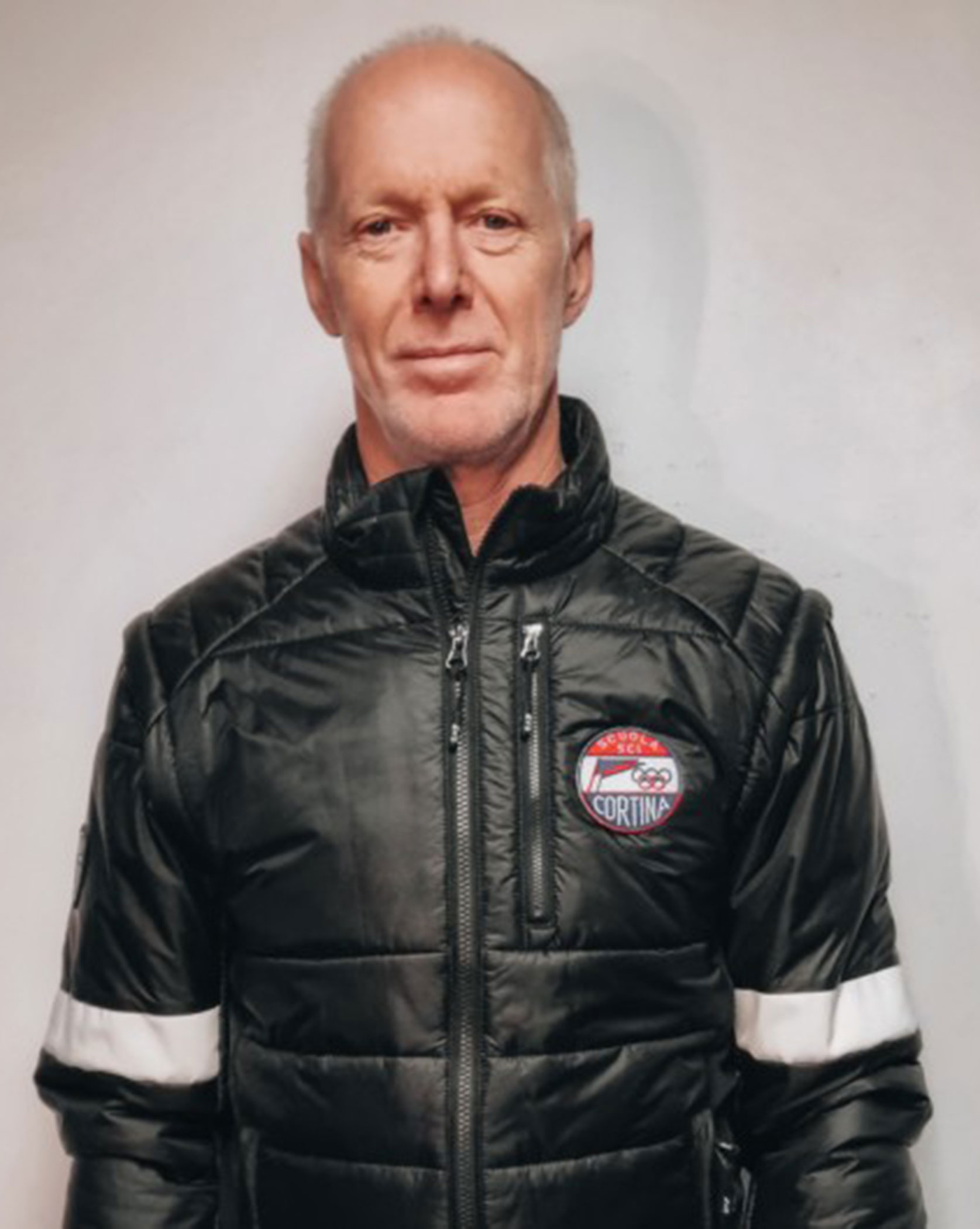 Man in a black jacket with a Cortina badge stands against a plain white background, looking at the camera. - Scuola Sci Cortina