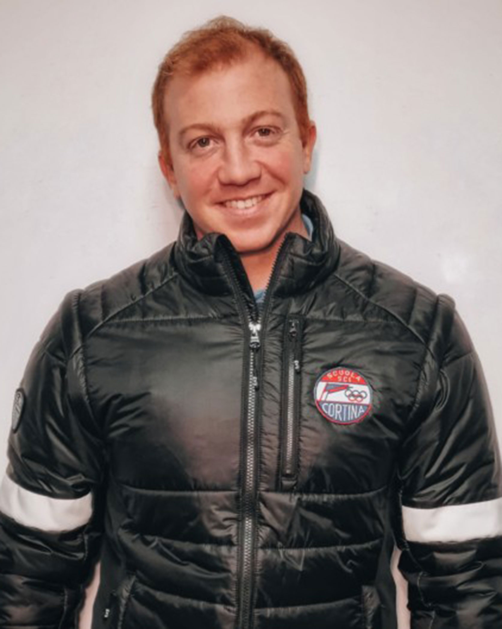Smiling person in a black puffer jacket with a Cortina patch, standing against a plain white wall. - Scuola Sci Cortina