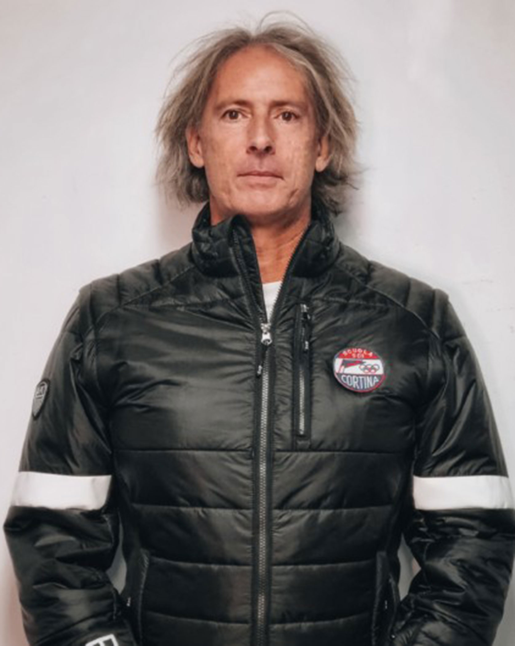 A man with gray hair wears a black puffer jacket with a Cortina patch, standing against a light background. - Scuola Sci Cortina