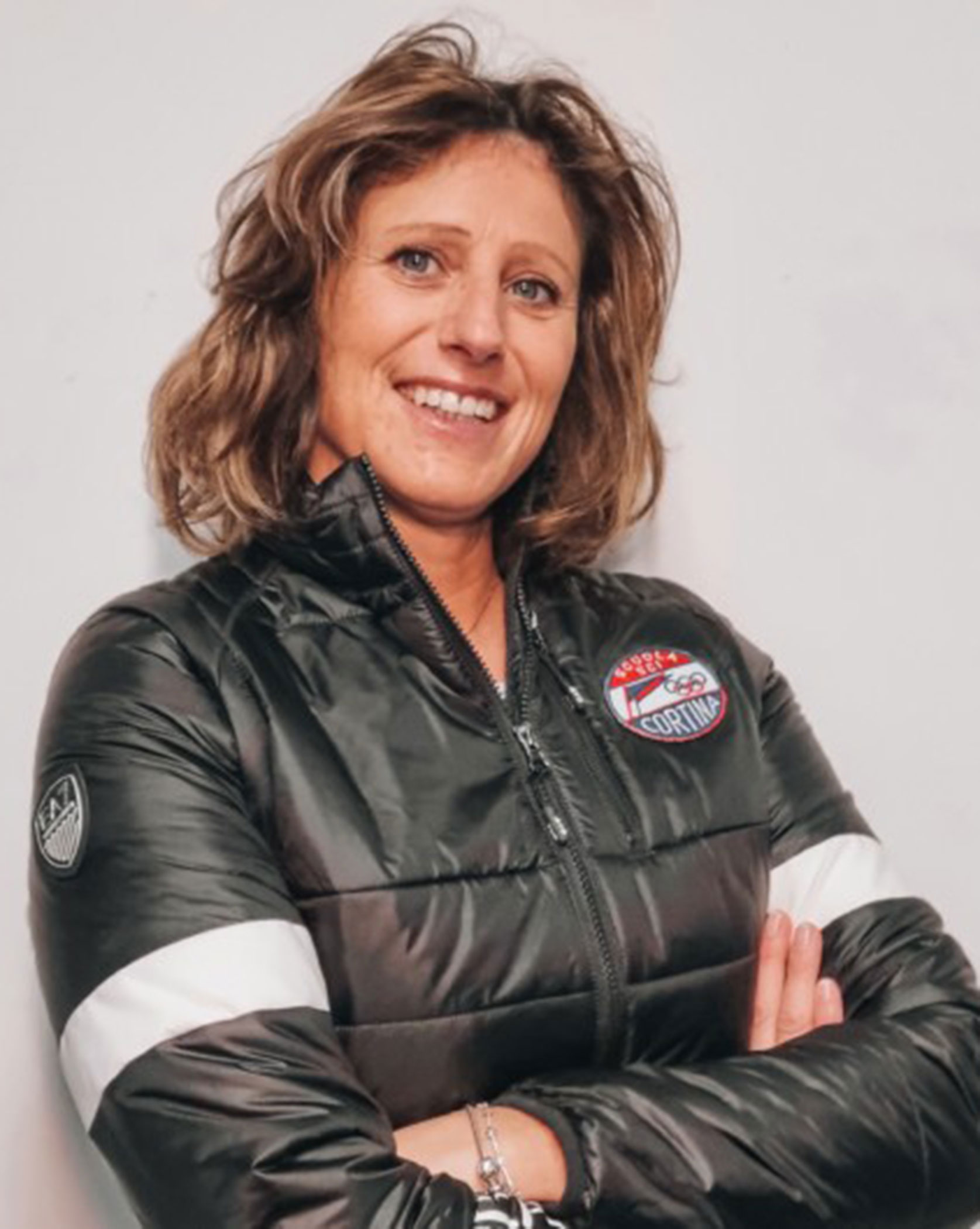 Woman in a black jacket with folded arms, smiling, standing against a plain white background. - Scuola Sci Cortina