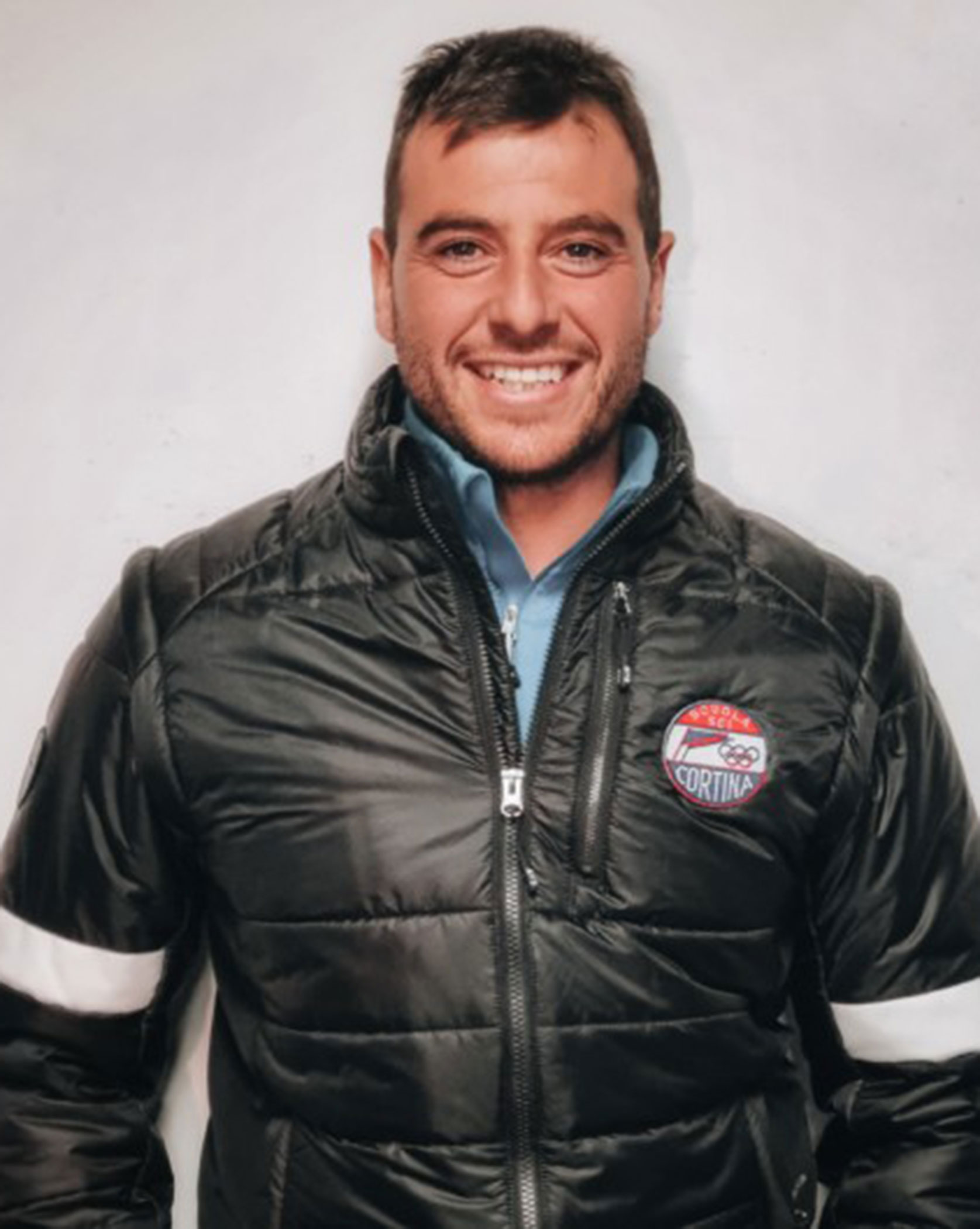 Smiling man in a black puffer jacket with a Cortina patch on the chest, standing against a light background. - Scuola Sci Cortina