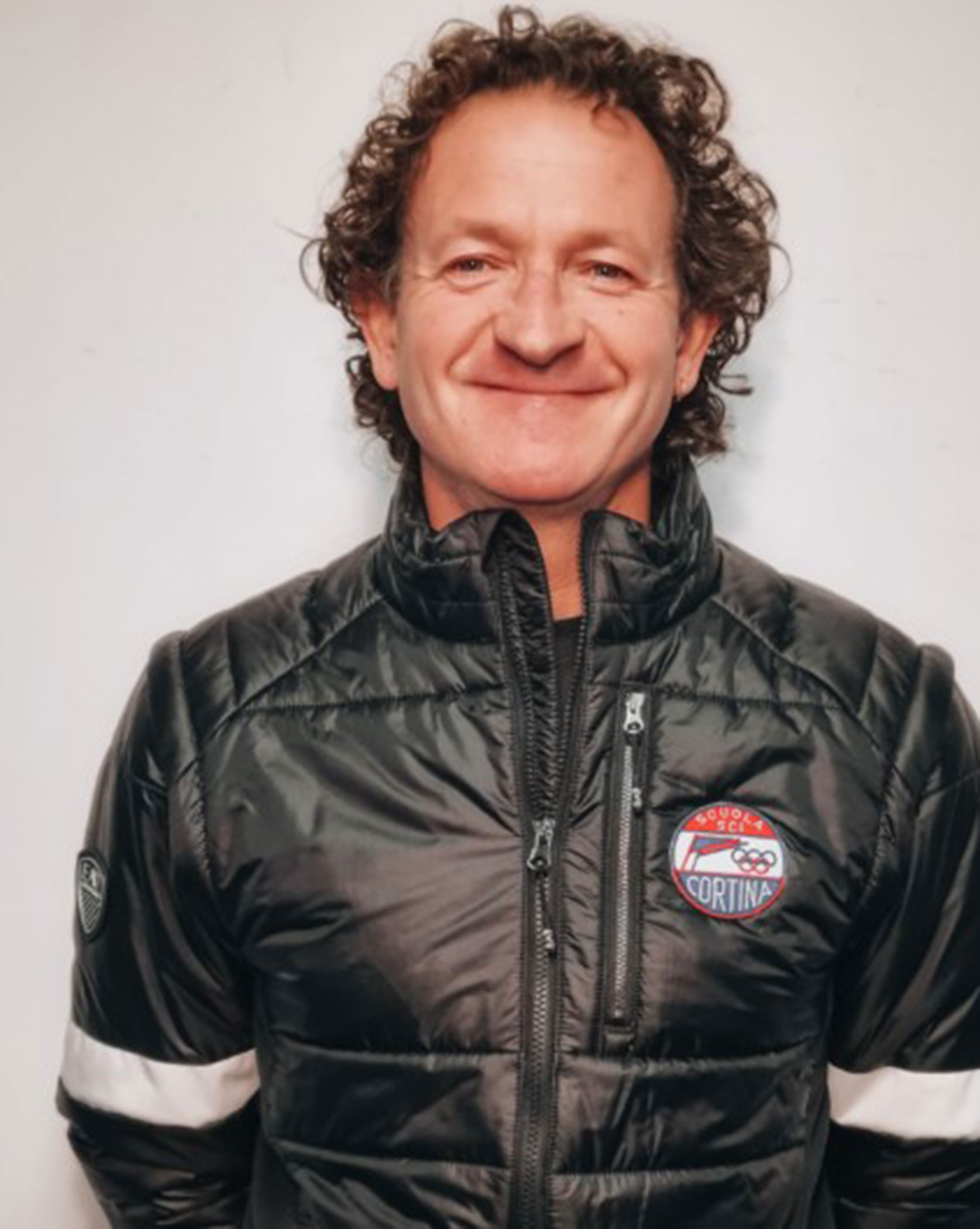 A man with curly hair smiles, wearing a black jacket with a Cortina logo, standing against a white wall. - Scuola Sci Cortina
