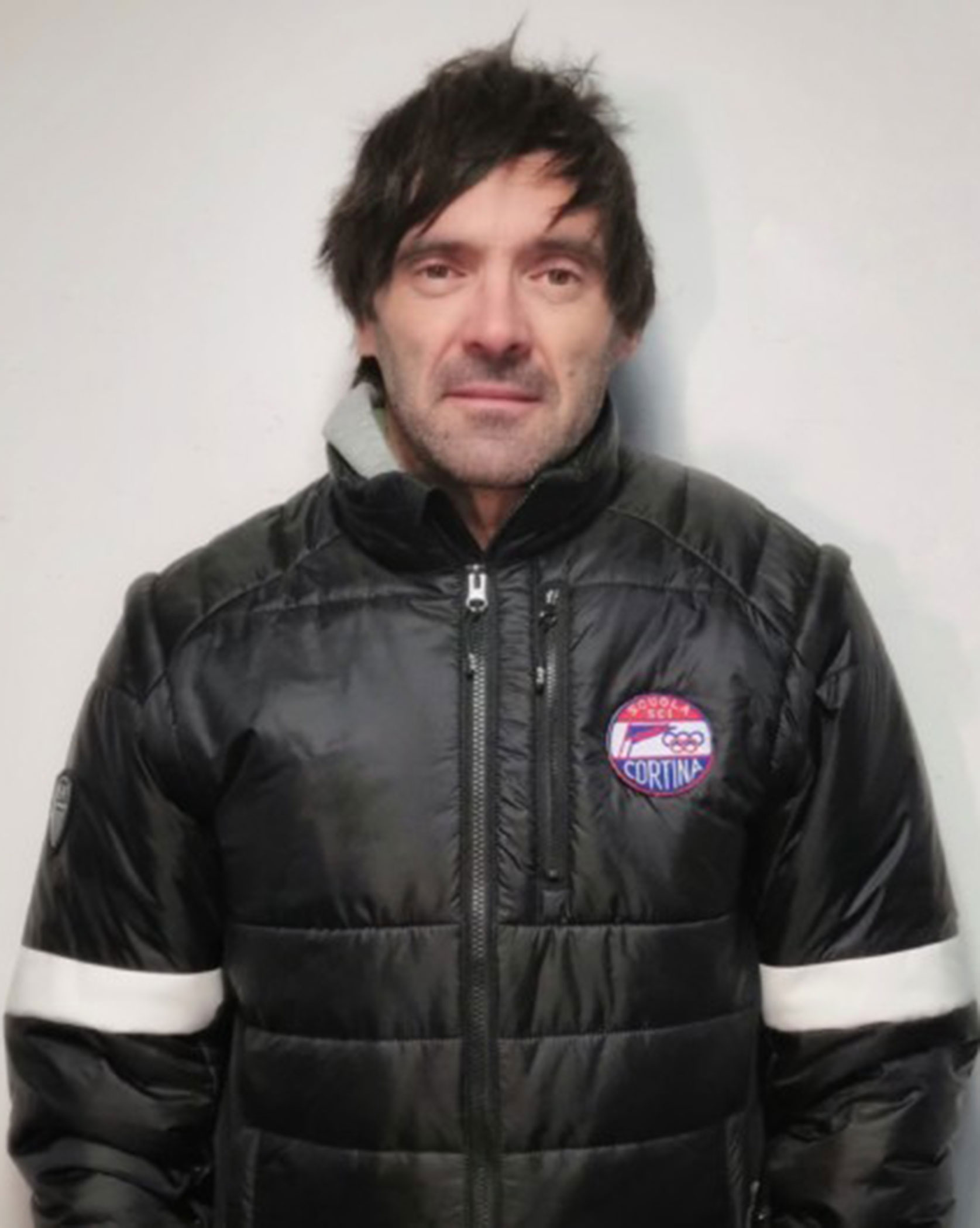 Man with dark hair wearing a black jacket with a Cortina patch, standing against a plain background. - Scuola Sci Cortina