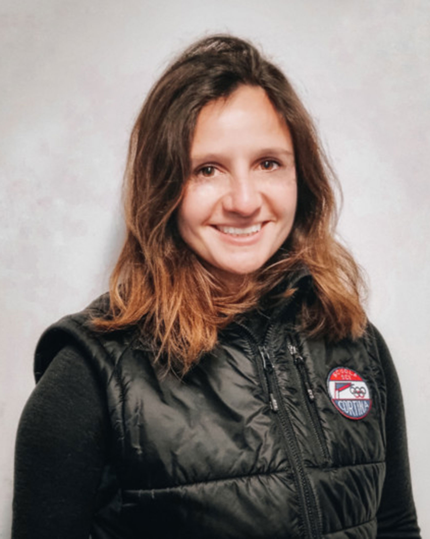 Woman with wavy brown hair, wearing a black vest with a Cortina patch, smiling at the camera. - Scuola Sci Cortina