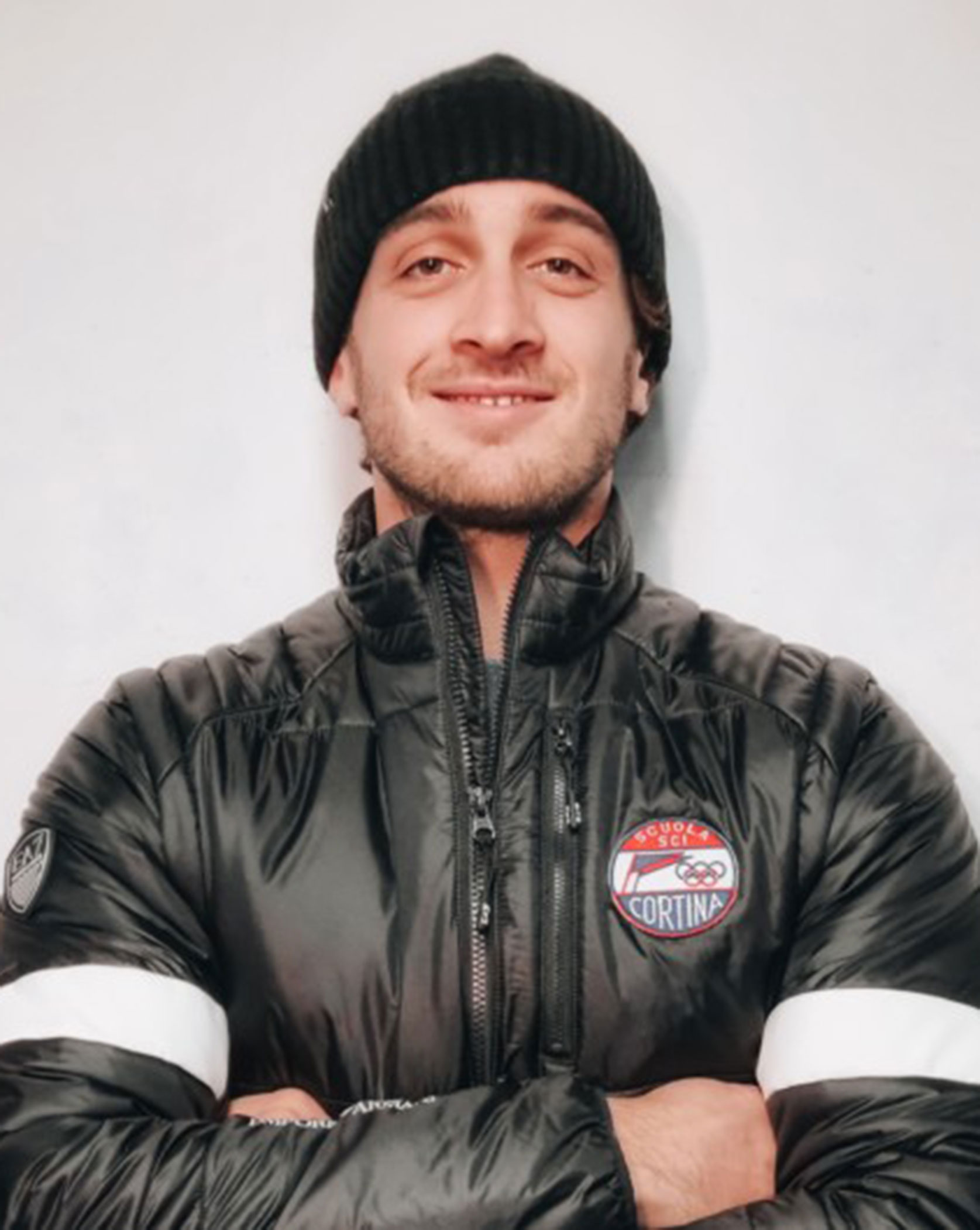 Man in a black jacket and beanie stands with arms crossed, smiling at the camera against a plain background. - Scuola Sci Cortina