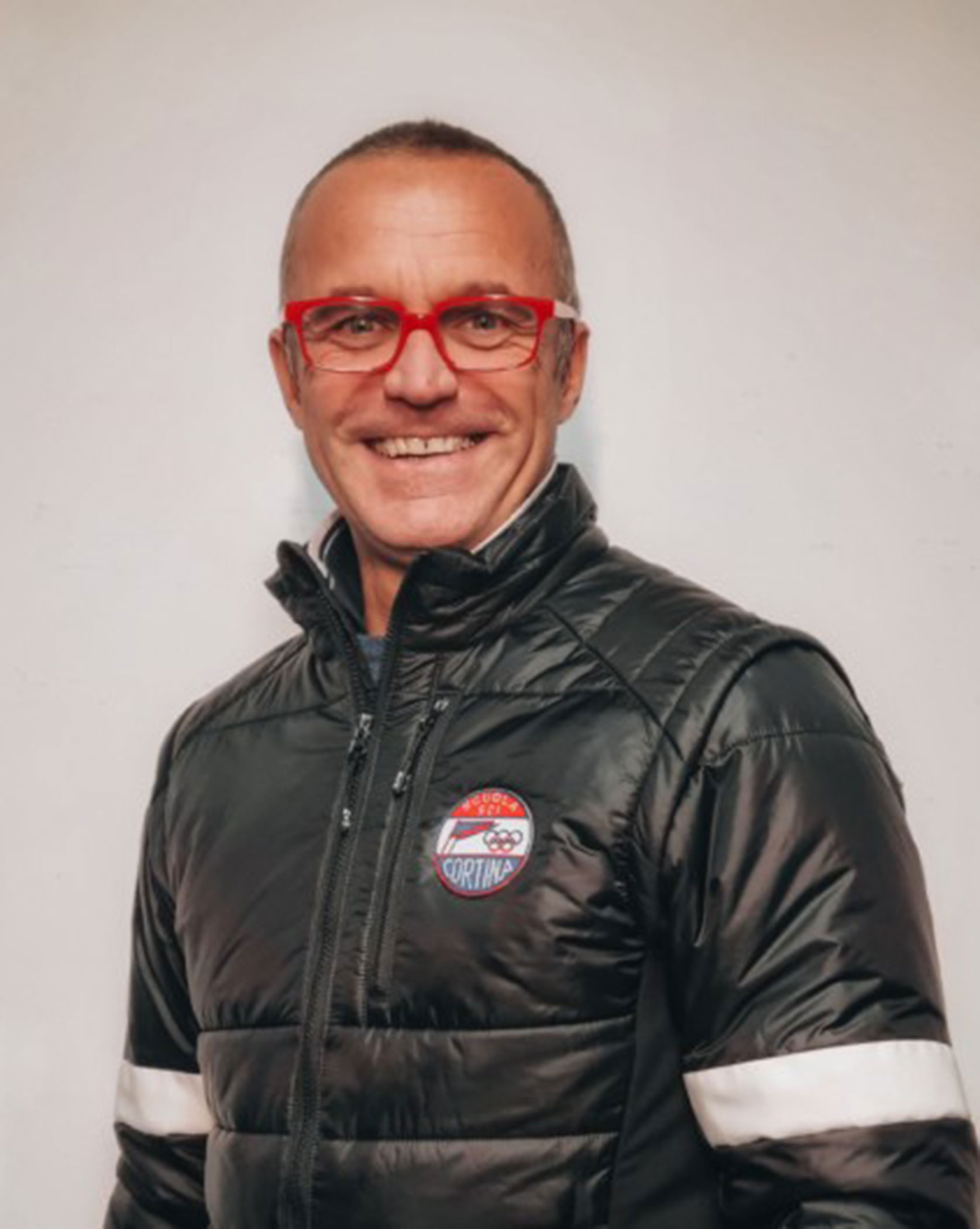 Smiling person in red glasses and a black puffer jacket with a Cortina logo, standing by a white wall. - Scuola Sci Cortina