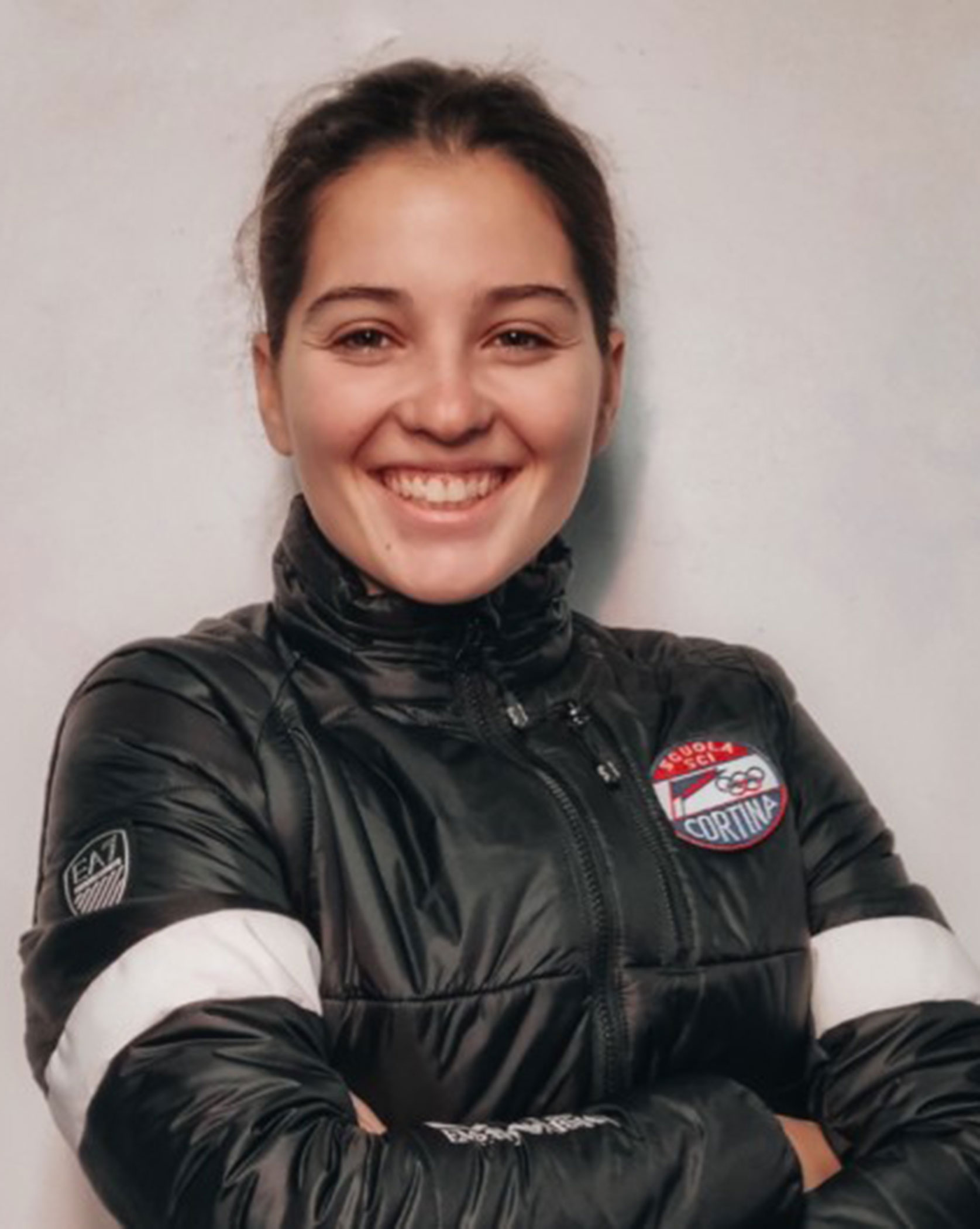 Woman in a black jacket with arms crossed, smiling in front of a light-colored background. - Scuola Sci Cortina