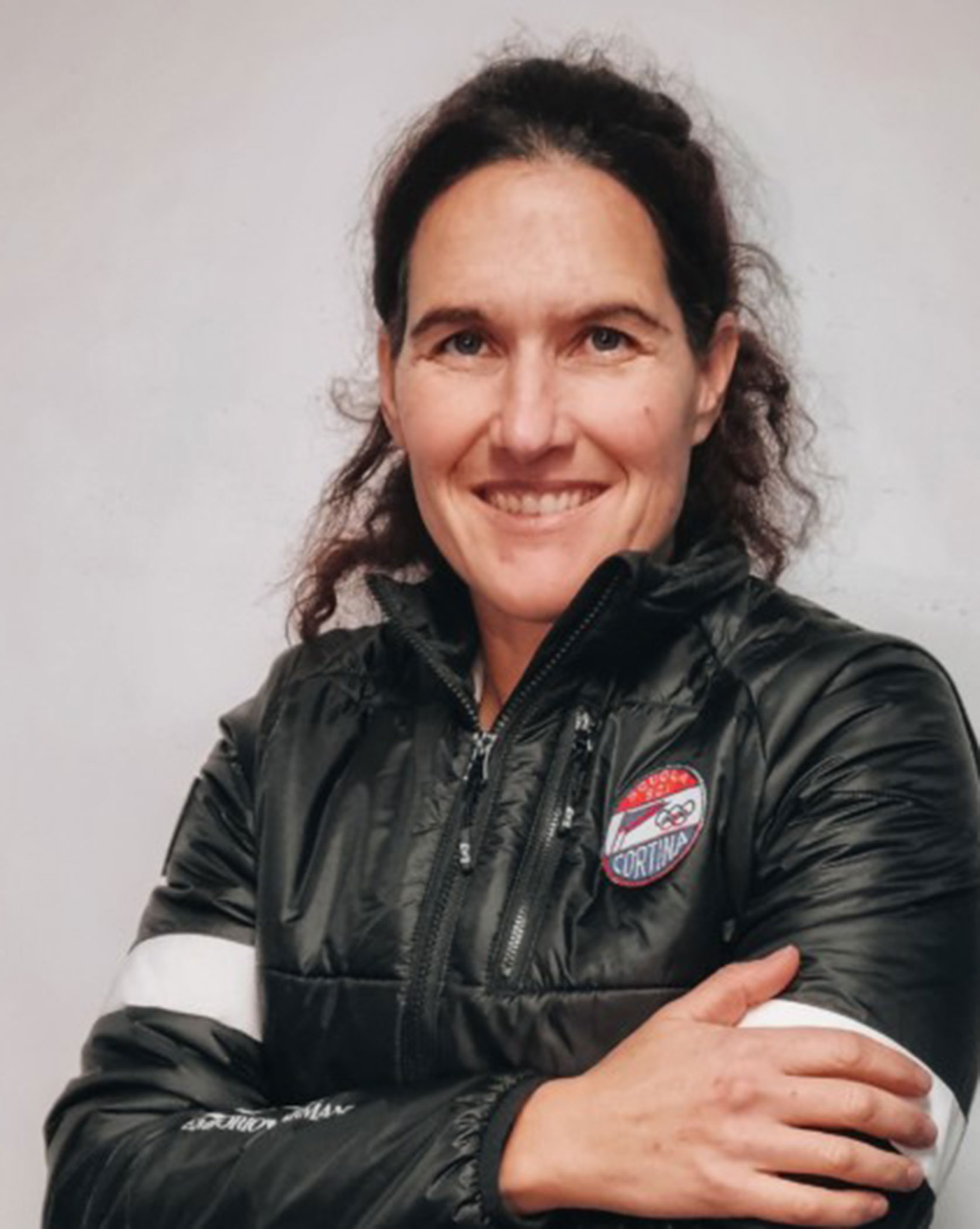 Woman with dark hair in a black jacket stands smiling with arms crossed against a plain background. - Scuola Sci Cortina