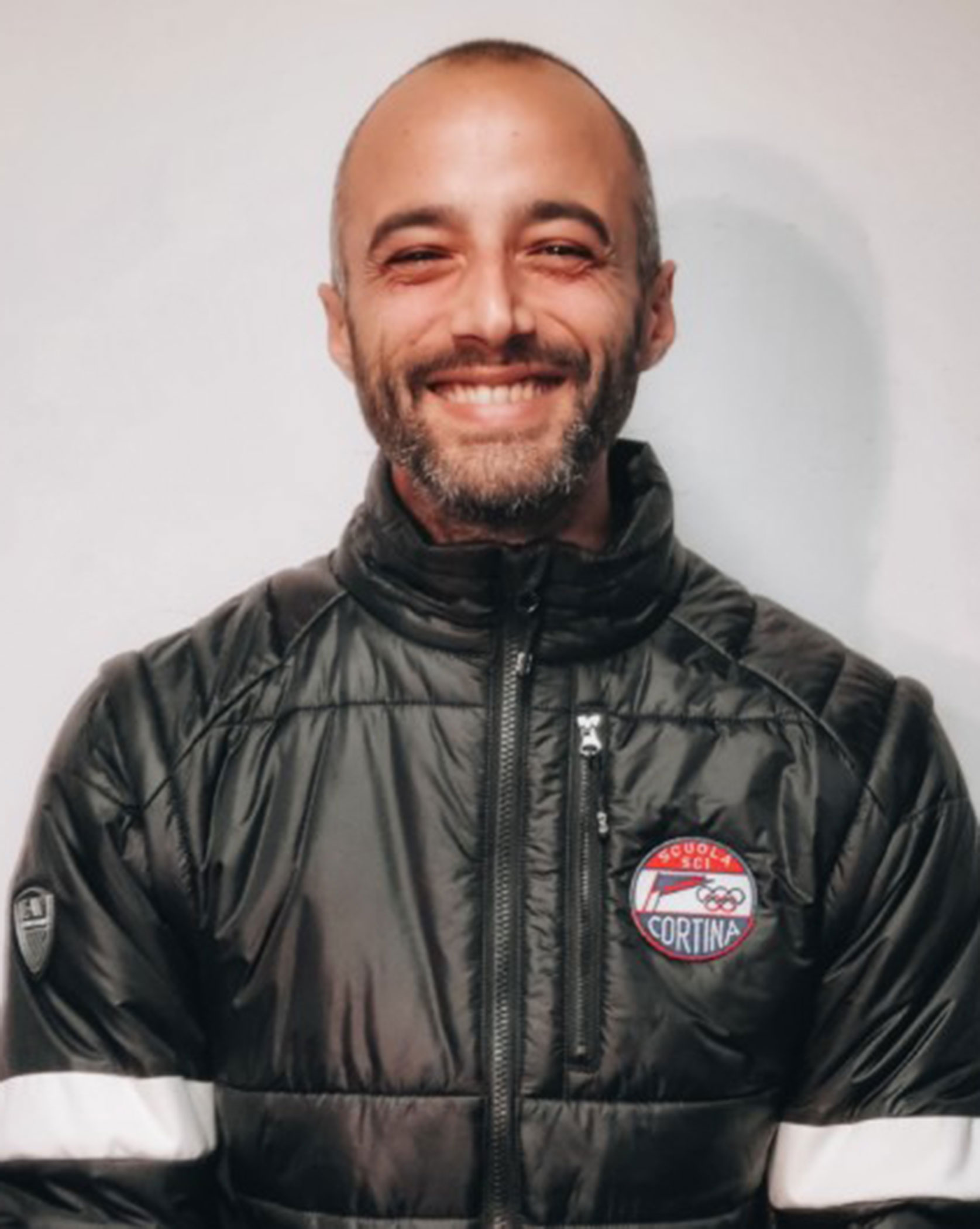 Smiling man with a beard in a black puffer jacket with a Cortina patch on the chest, against a light background. - Scuola Sci Cortina