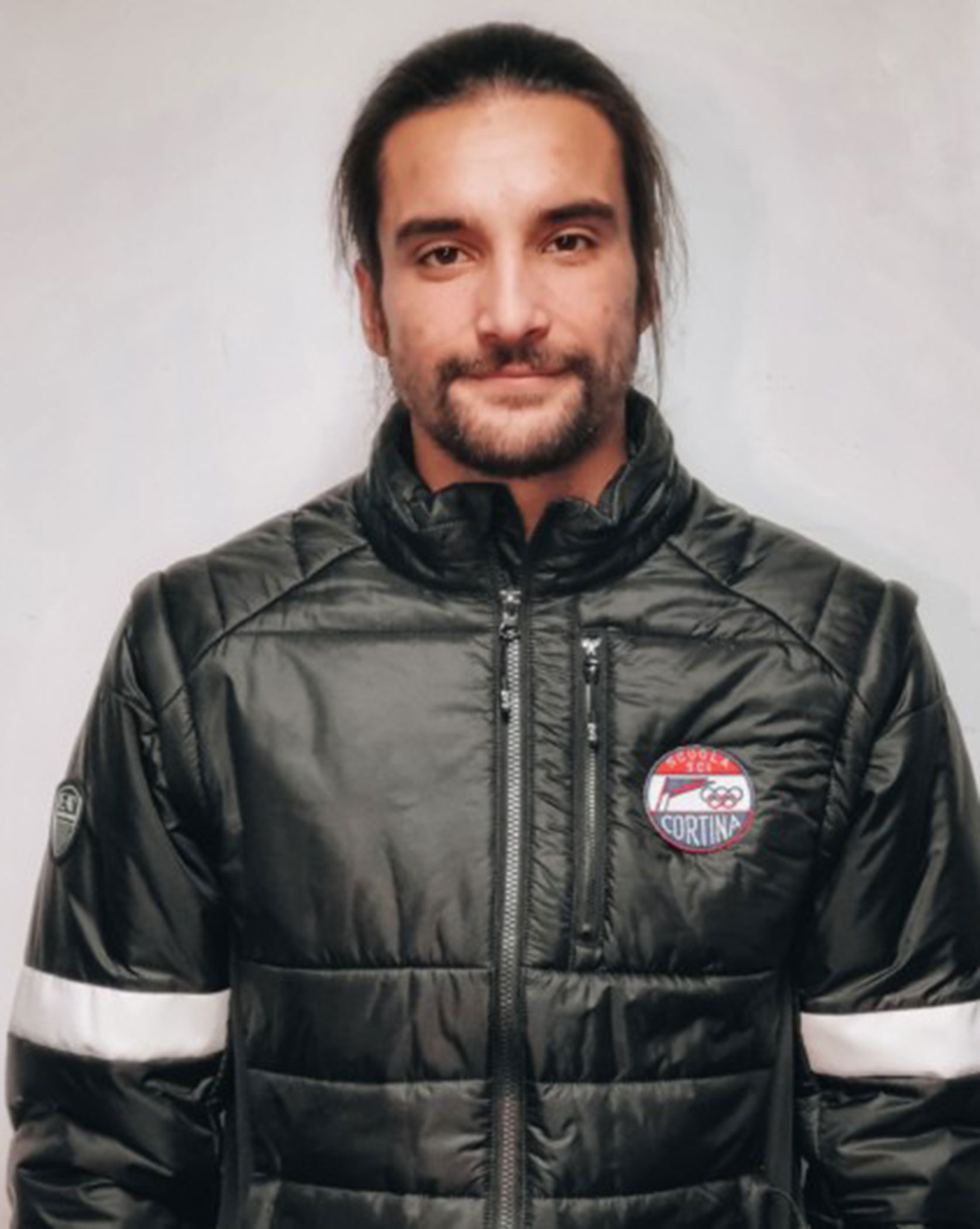 Man with long hair and beard wearing a black jacket with a Cortina patch, standing against a plain background. - Scuola Sci Cortina