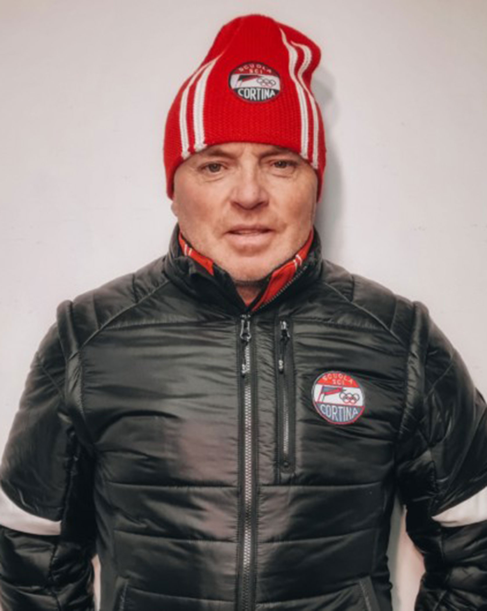 Person in a black jacket and red striped beanie with a Cortina logo, standing against a plain background. - Scuola Sci Cortina