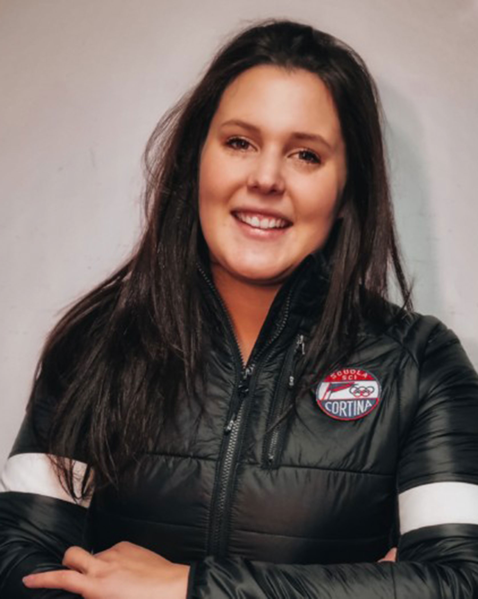 Woman with long dark hair smiles, wearing a black jacket with a Cortina badge on the chest. - Scuola Sci Cortina
