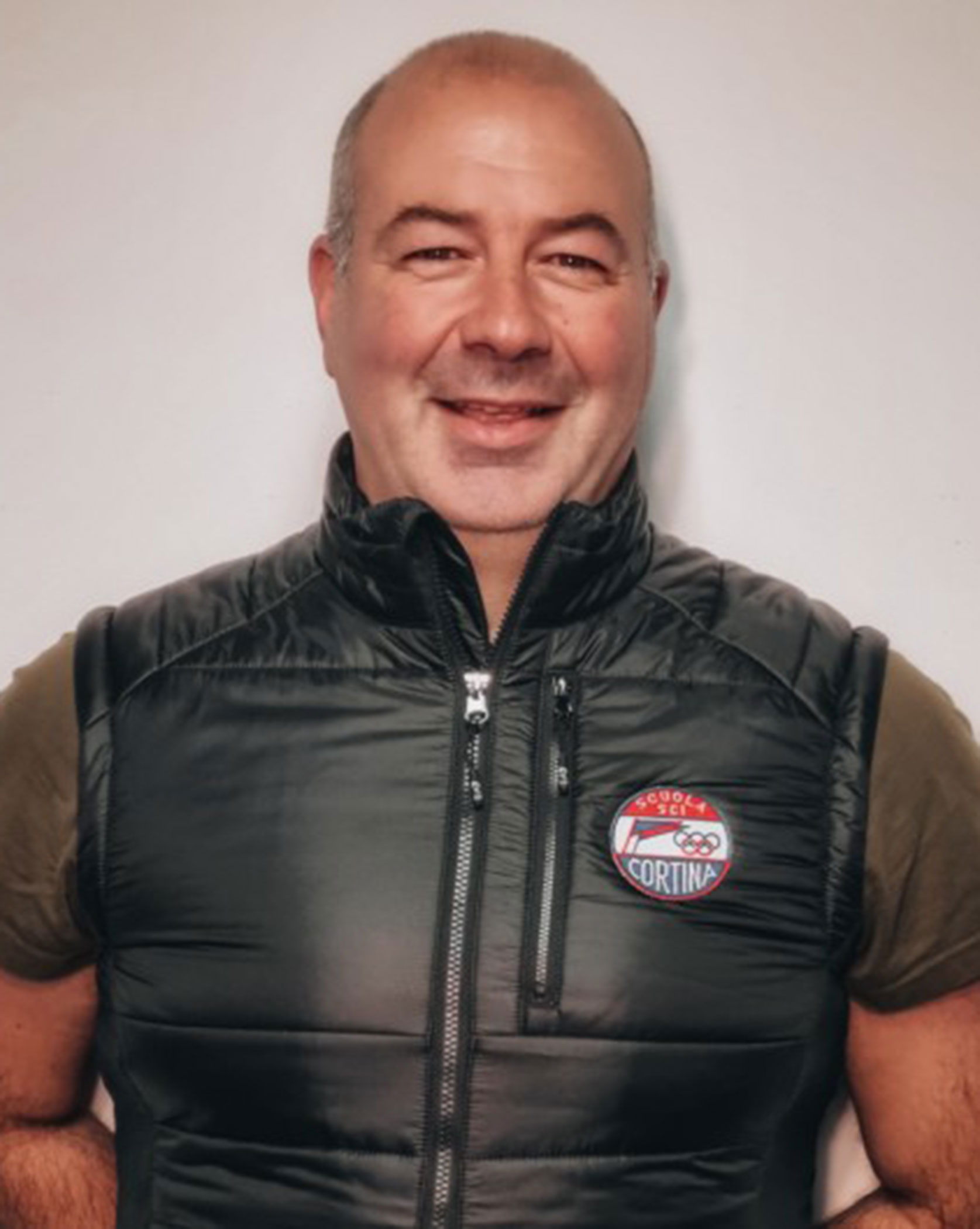 Smiling man in a puffer vest with an Equolo Cortina logo stands against a plain light background. - Scuola Sci Cortina