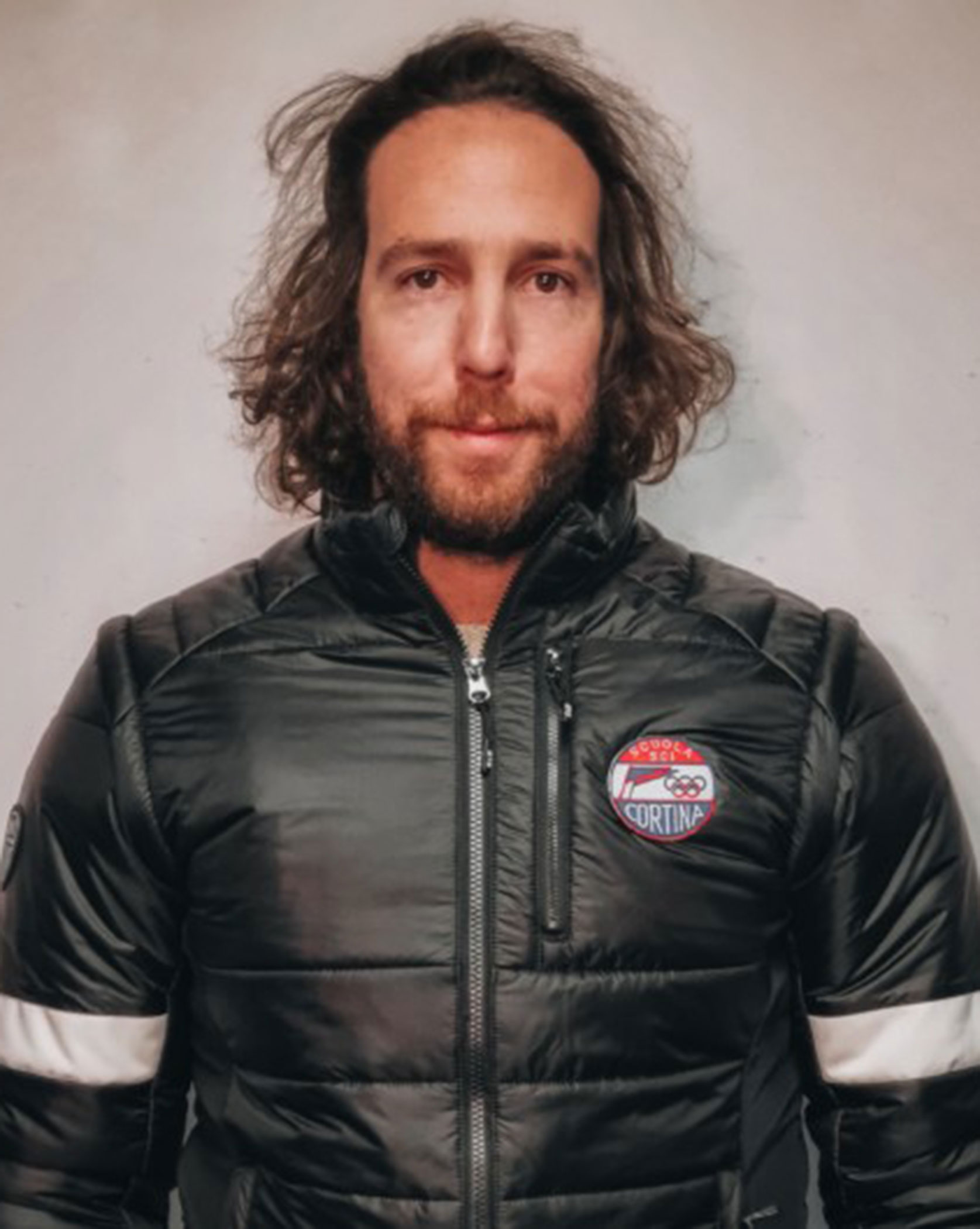 Man with long hair and beard wearing a black puffer jacket with a Cortina patch, standing against a wall. - Scuola Sci Cortina