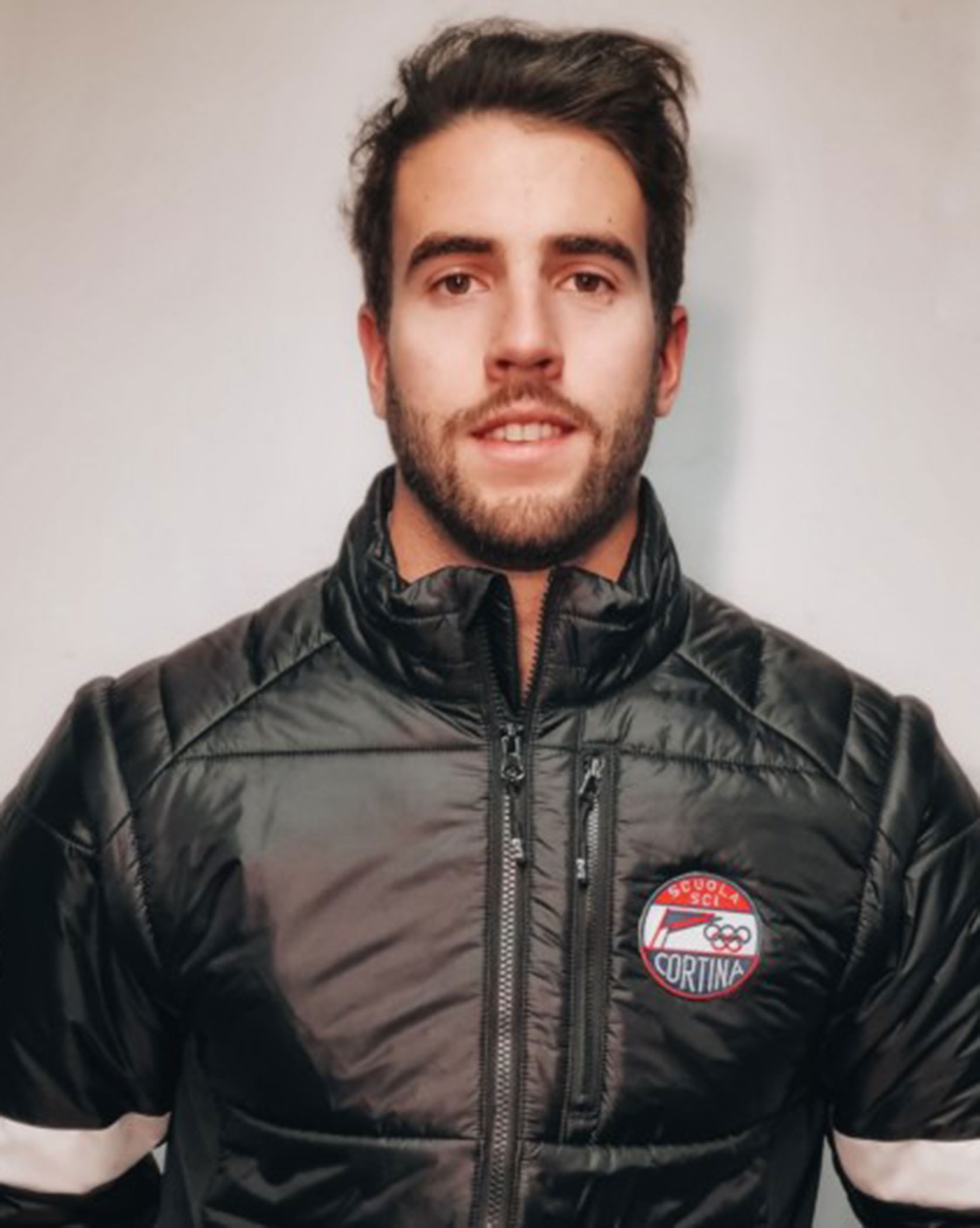 Man with dark hair in a black Cortina jacket stands against a plain light background, looking at the camera. - Scuola Sci Cortina