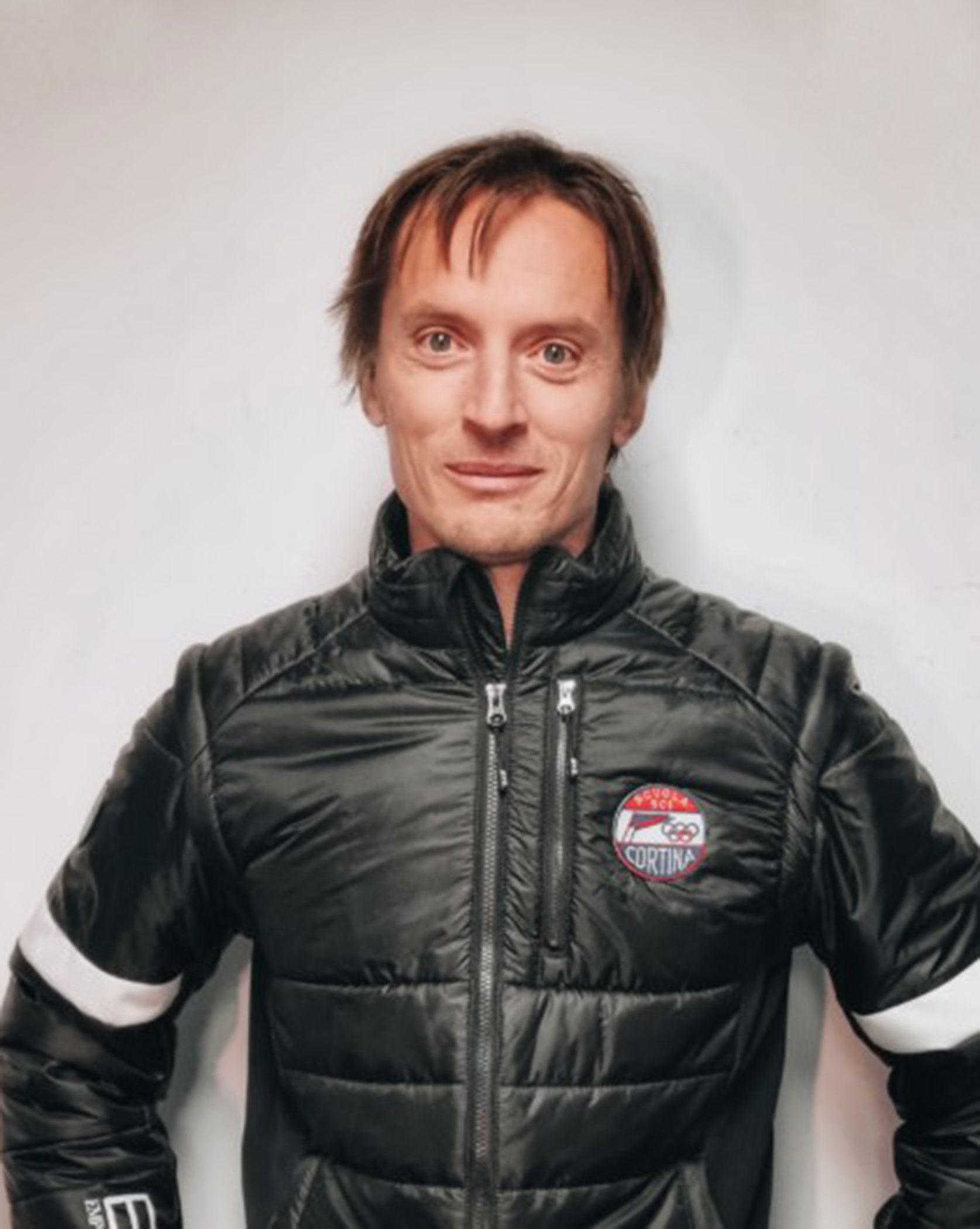 A person wearing a black jacket with a Cortina badge stands against a plain white background. - Scuola Sci Cortina