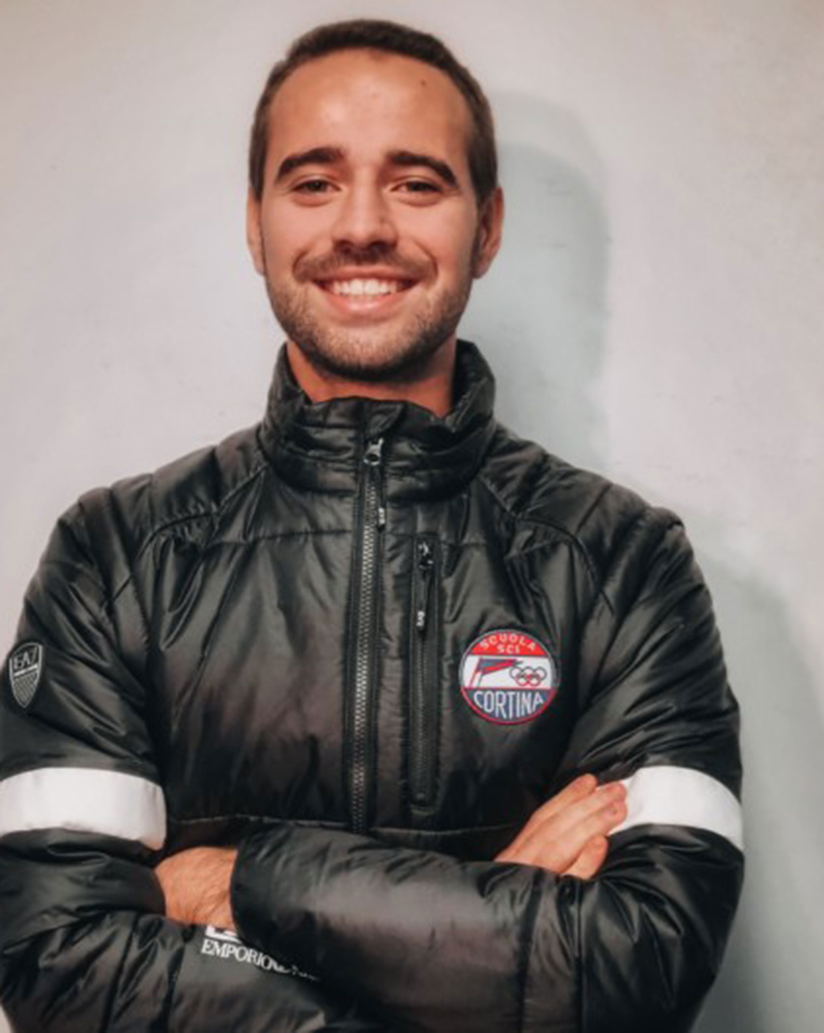 Man in a black jacket with crossed arms, smiling in front of a plain light-colored background. - Scuola Sci Cortina