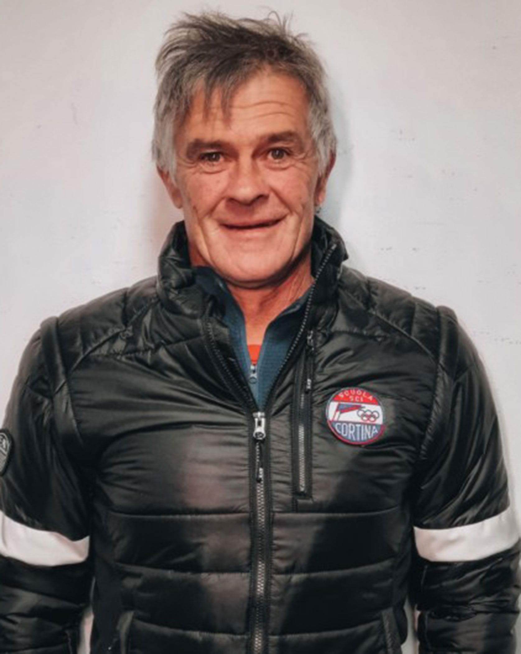 Smiling man in a black puffer jacket with a Cortina logo, standing against a plain white wall. - Scuola Sci Cortina