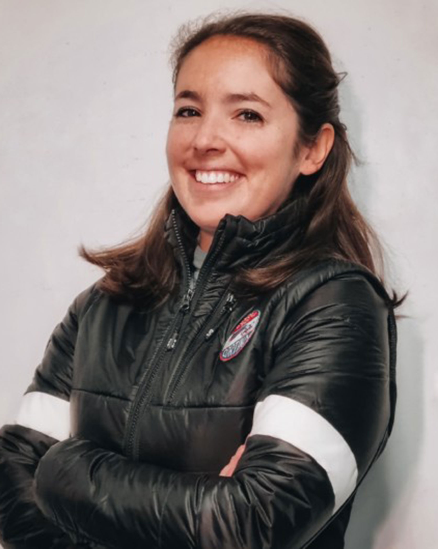 Woman in a black puffer jacket with arms crossed, smiling in front of a plain light-colored wall. - Scuola Sci Cortina