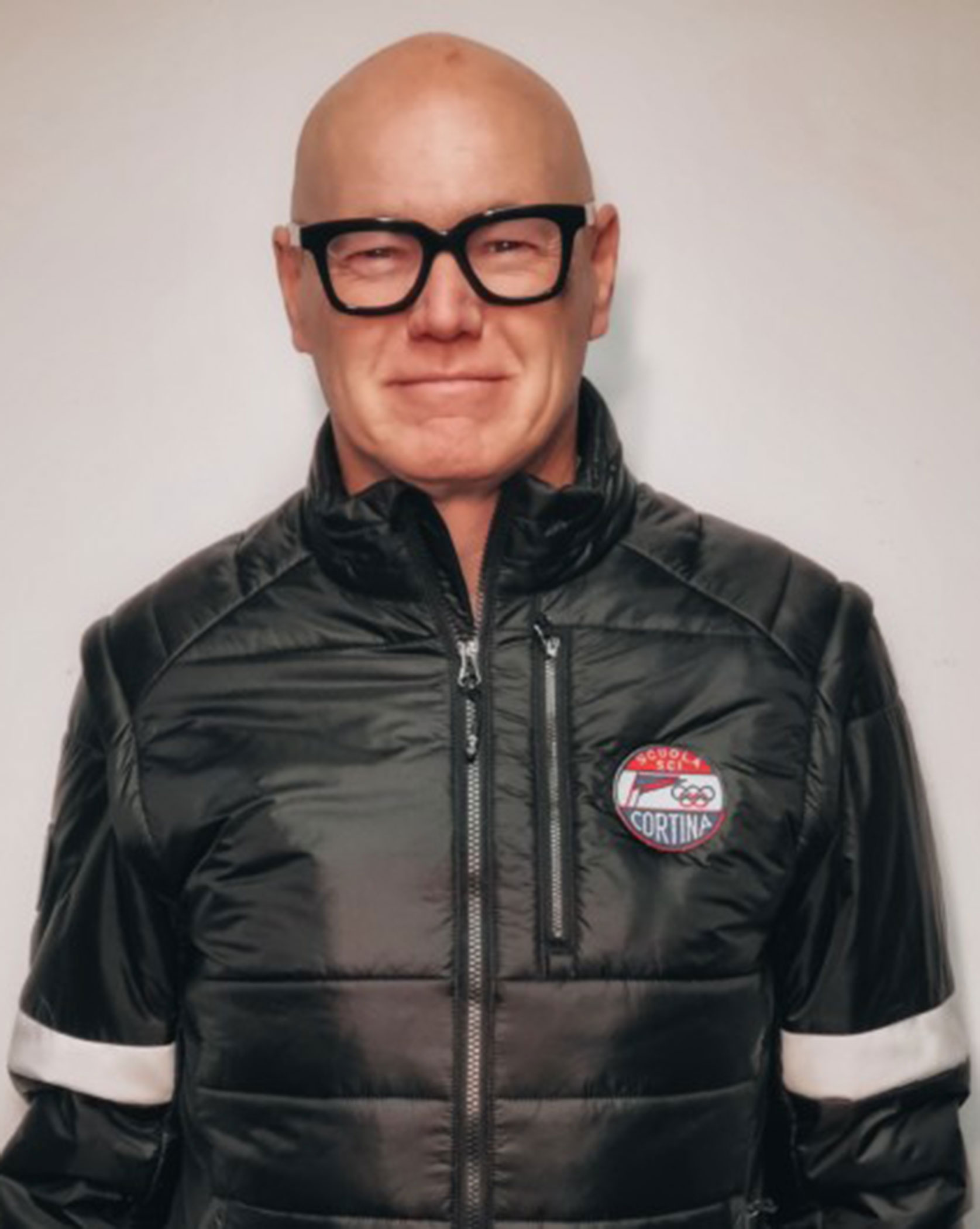 A person in glasses and a black jacket with a Cortina patch, standing against a plain background. - Scuola Sci Cortina