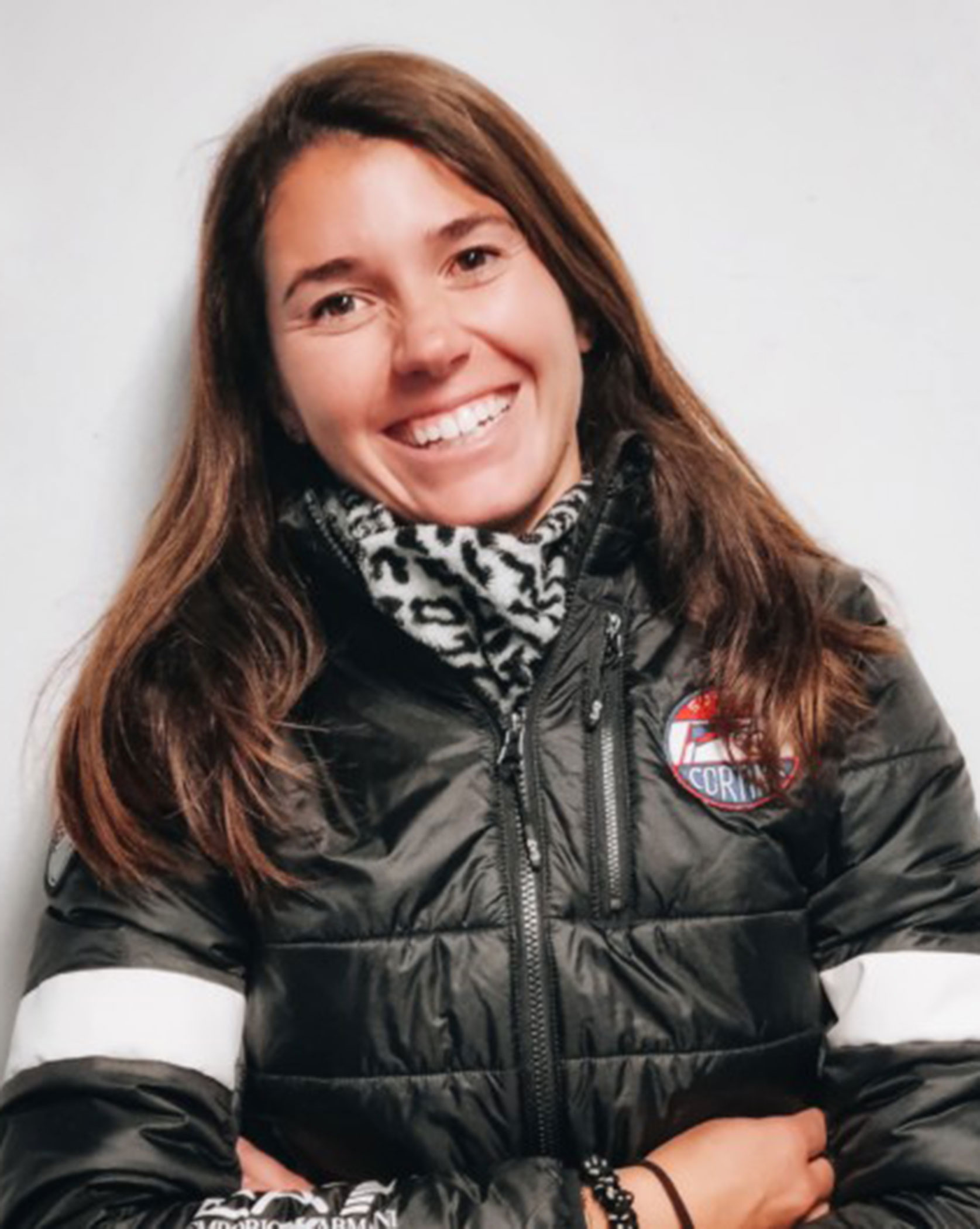 Smiling woman in a black jacket with a logo, standing against a plain white wall. - Scuola Sci Cortina