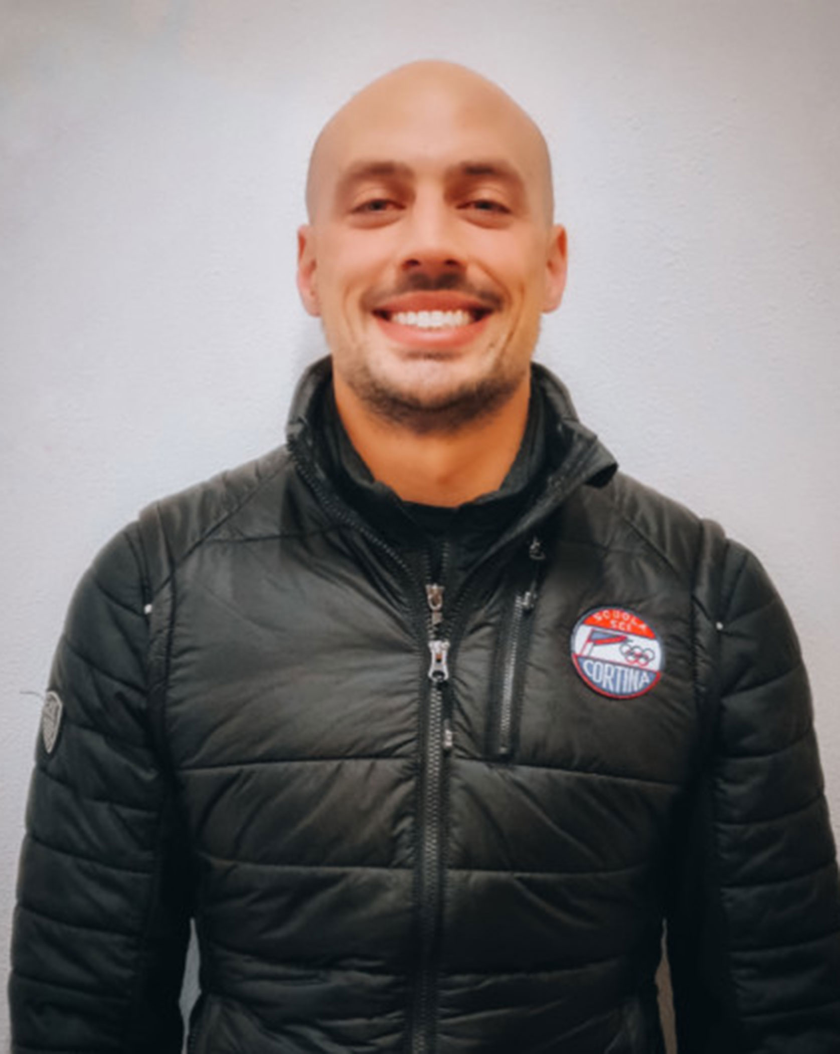 Smiling man in a black quilted jacket with a Cortina patch, standing against a plain light wall. - Scuola Sci Cortina