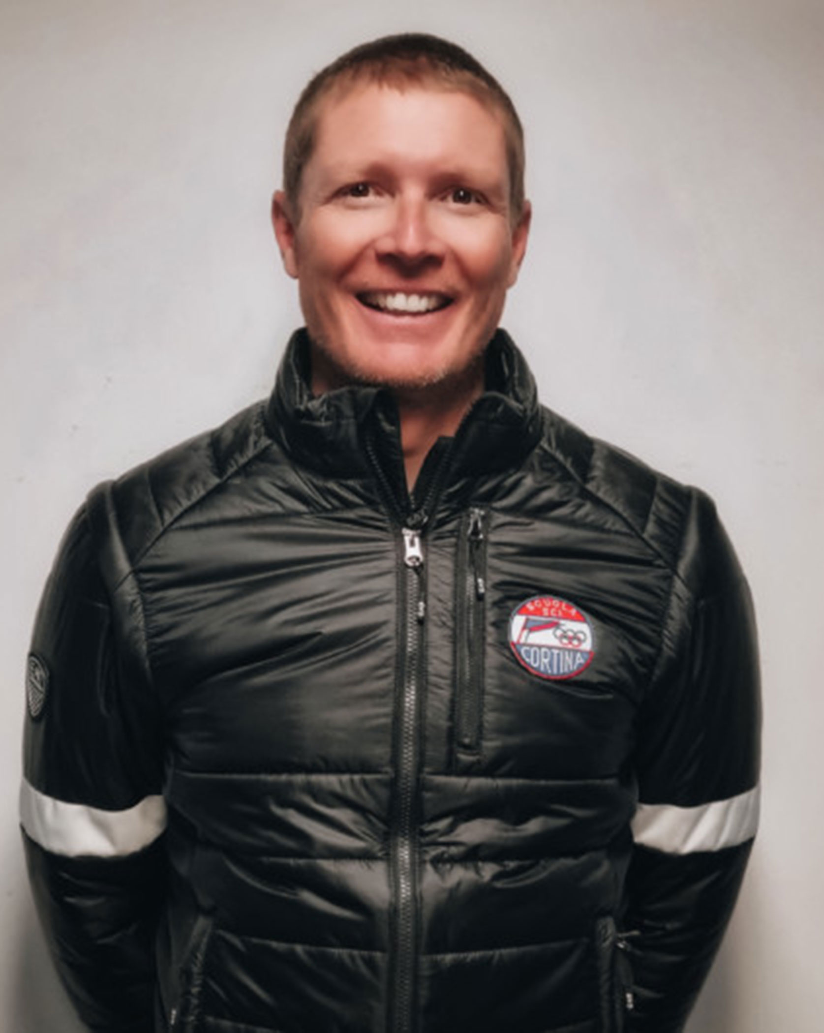 Smiling man in a black jacket with white stripes and a patch that reads Milano Cortina.. - Scuola Sci Cortina