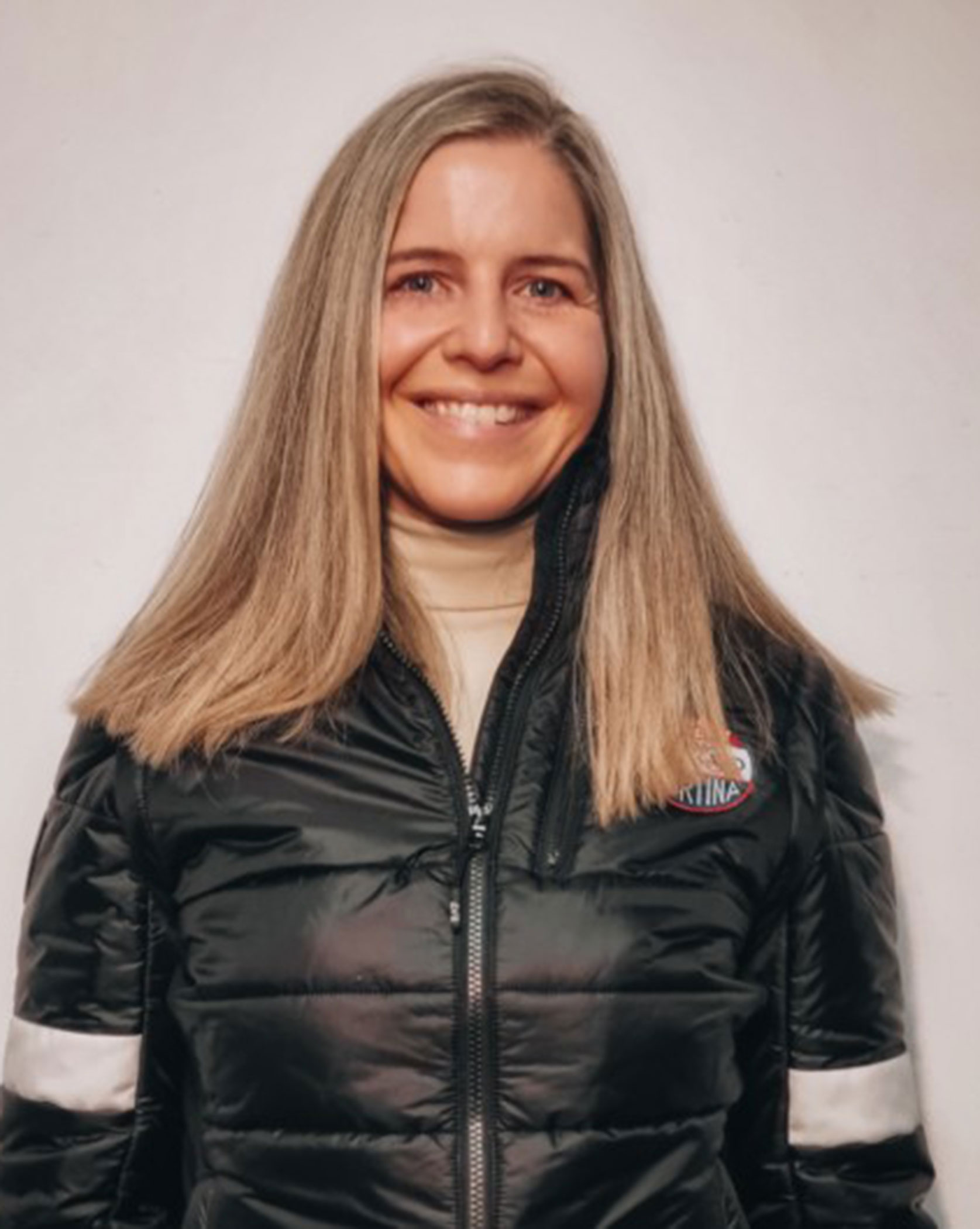 A woman with straight, shoulder-length hair smiles, wearing a black puffer jacket with a patch on the chest. - Scuola Sci Cortina