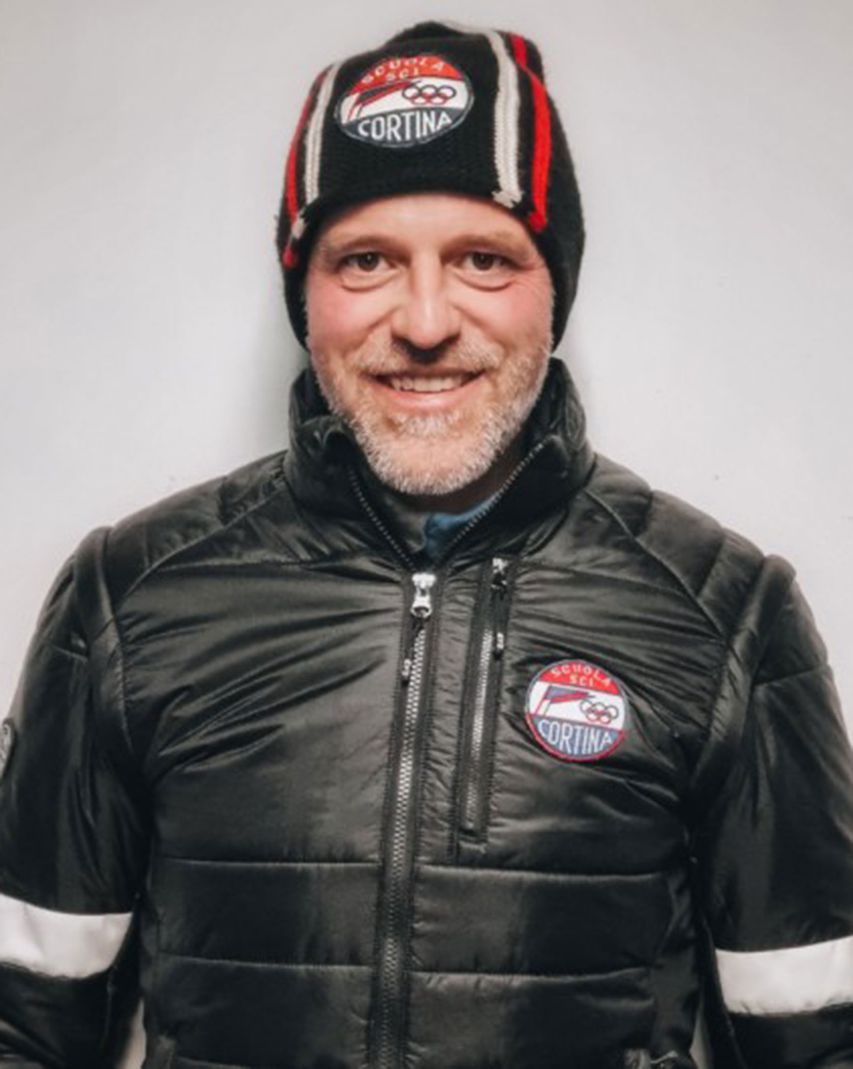 Smiling man in a black jacket and beanie with Cortina patches on a plain white background. - Scuola Sci Cortina