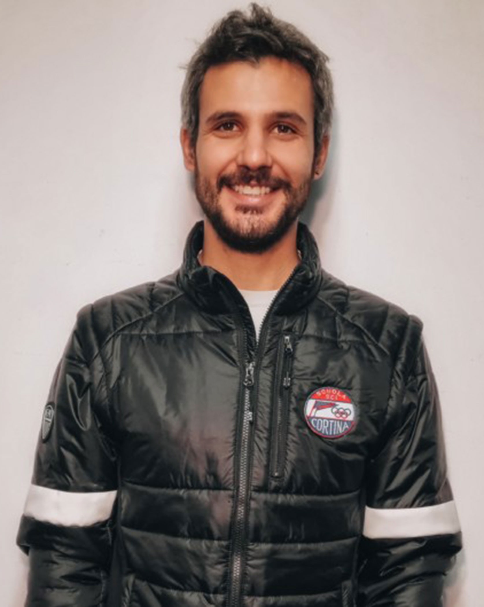 Man with short dark hair and beard smiles, wearing a black puffer jacket with a Cortina badge. - Scuola Sci Cortina