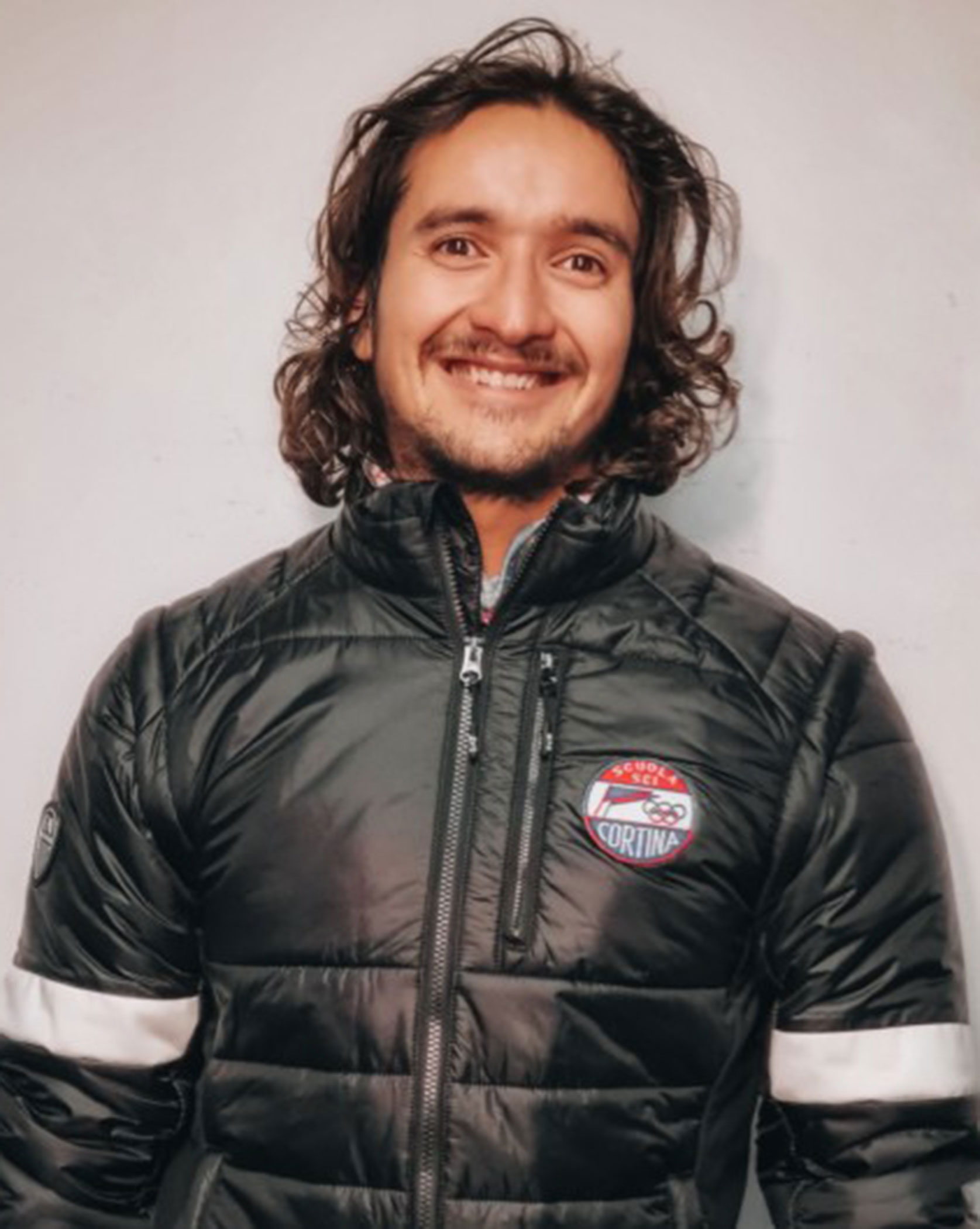 Smiling person with long hair in a black puffer jacket stands against a plain light-colored background. - Scuola Sci Cortina