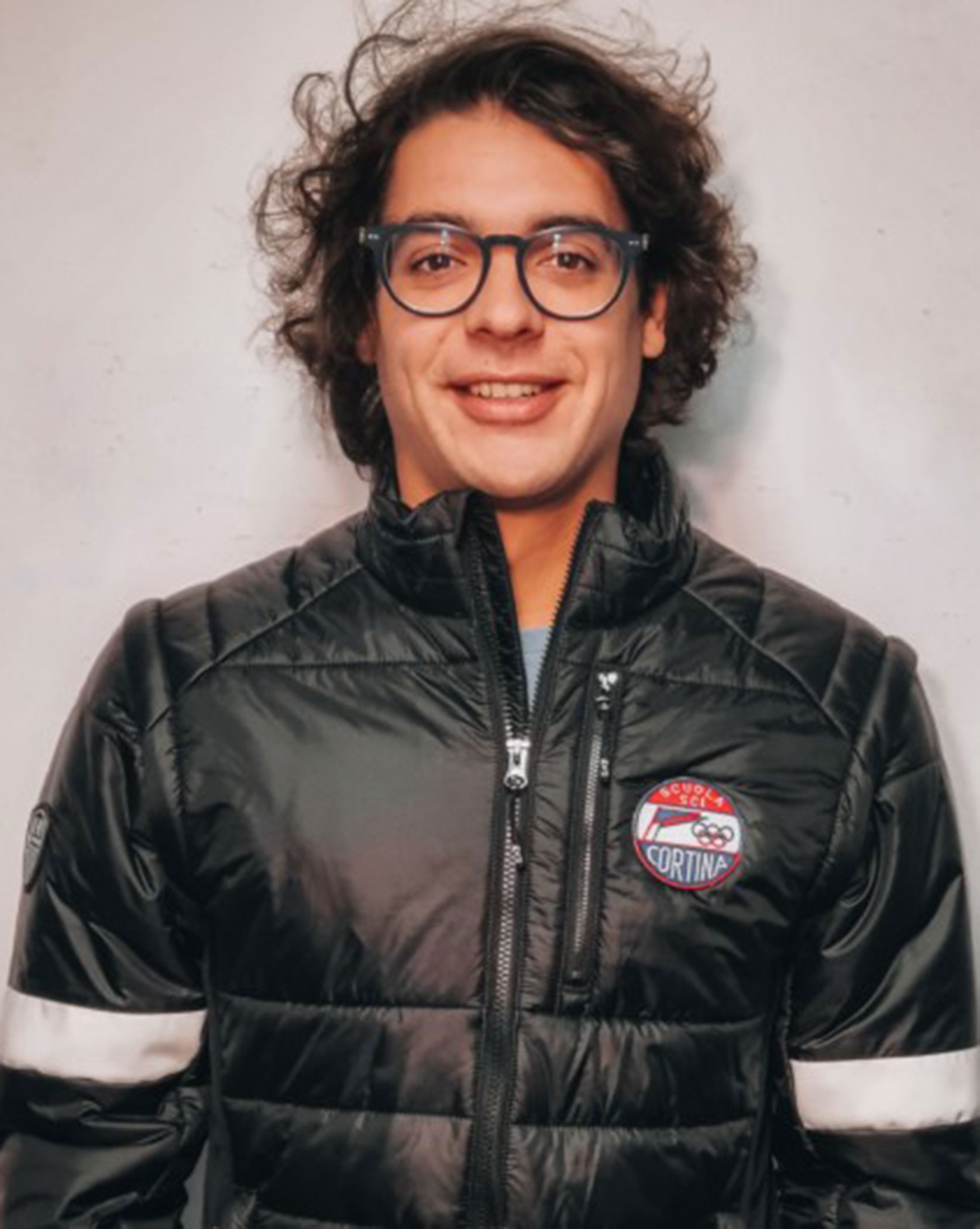 Person with curly hair and glasses, smiling, wearing a black puffer jacket with a Cortina logo badge. - Scuola Sci Cortina