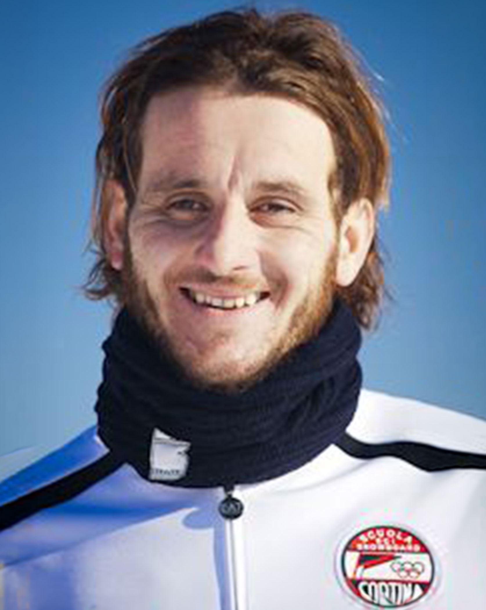 A man with brown hair and a beard, wearing a white jacket and black neck warmer, smiling outdoors. - Scuola Sci Cortina