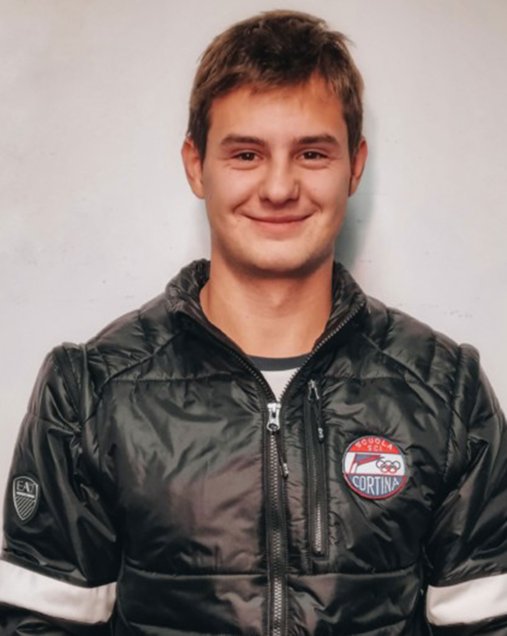 A young man in a black jacket with a Cortina logo smiles in front of a plain light background. - Scuola Sci Cortina