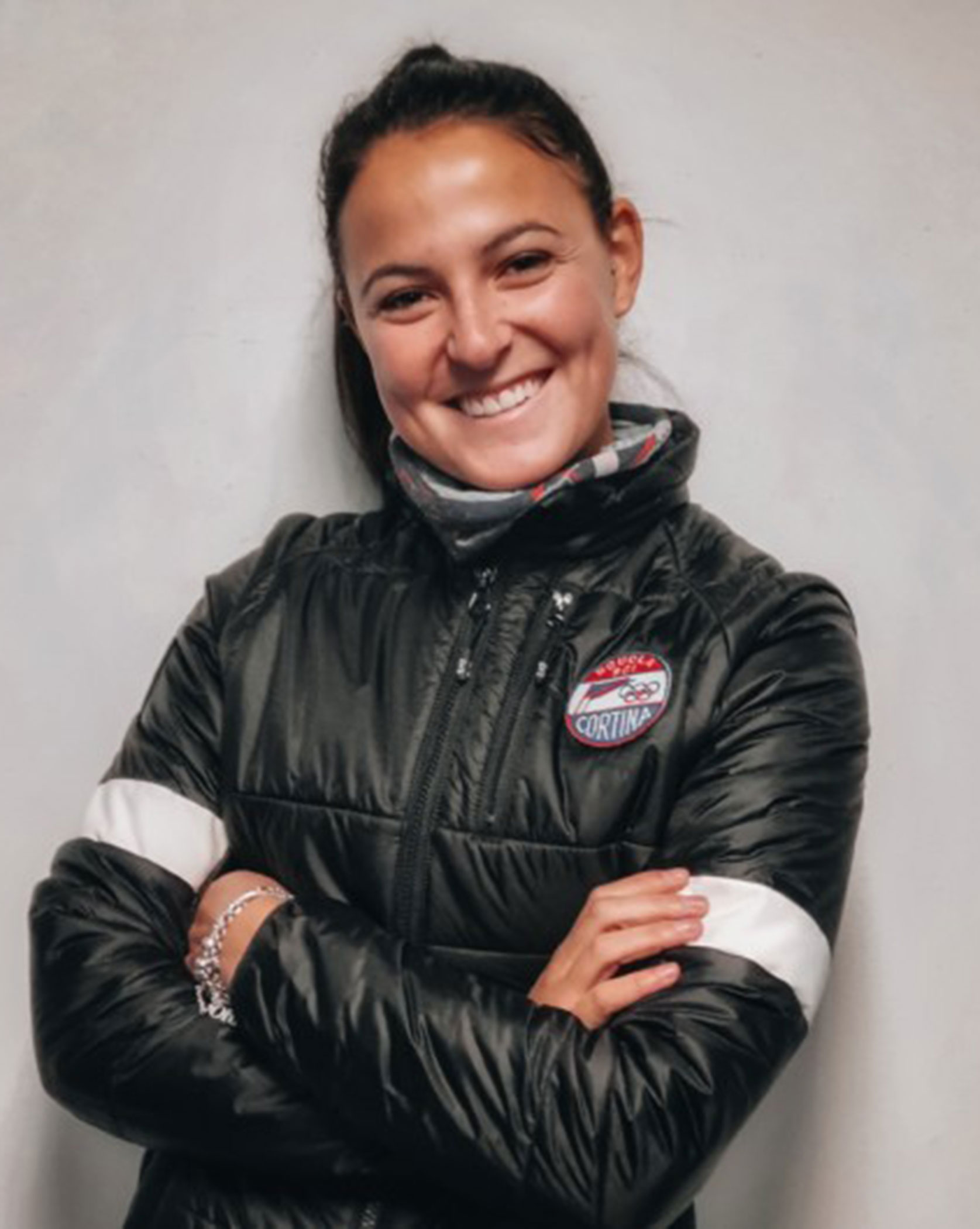Woman smiling with arms crossed, wearing a black jacket with a Cortina patch, standing against a light wall. - Scuola Sci Cortina