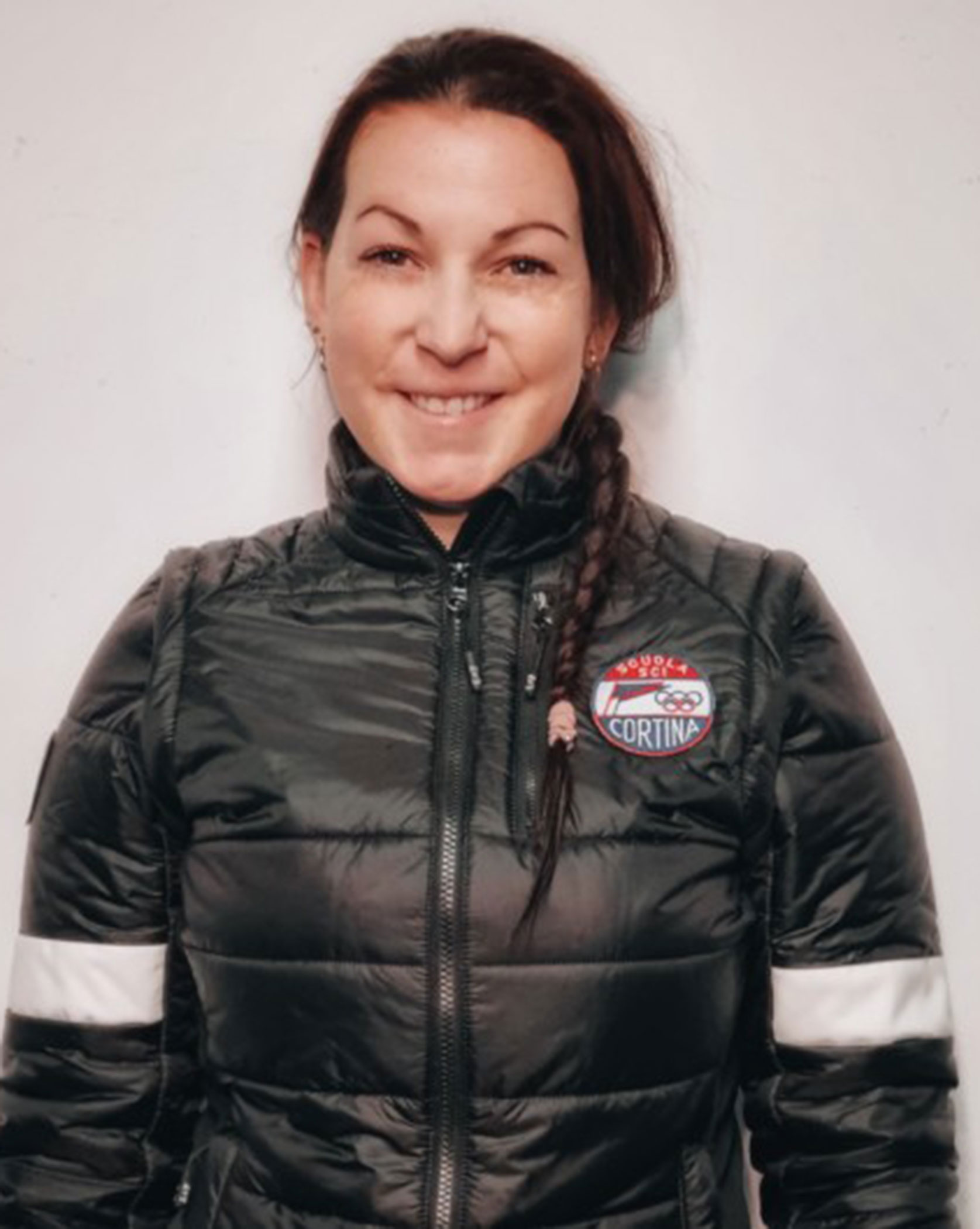 Woman with a braid smiling, wearing a black puffer jacket with a Cortina patch on the left side. - Scuola Sci Cortina