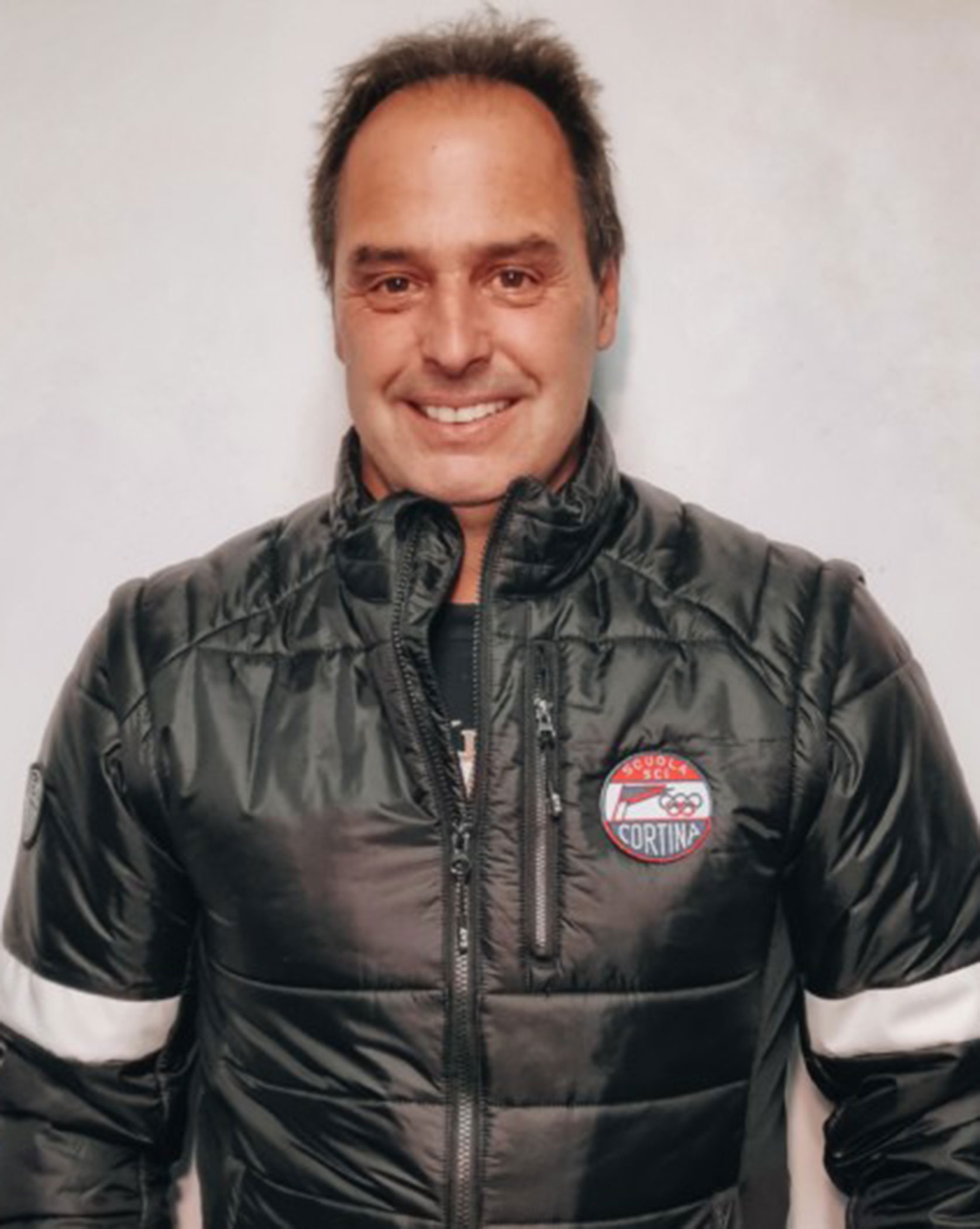 Smiling man in a dark puffer jacket with a Cortina badge, standing against a plain light background. - Scuola Sci Cortina