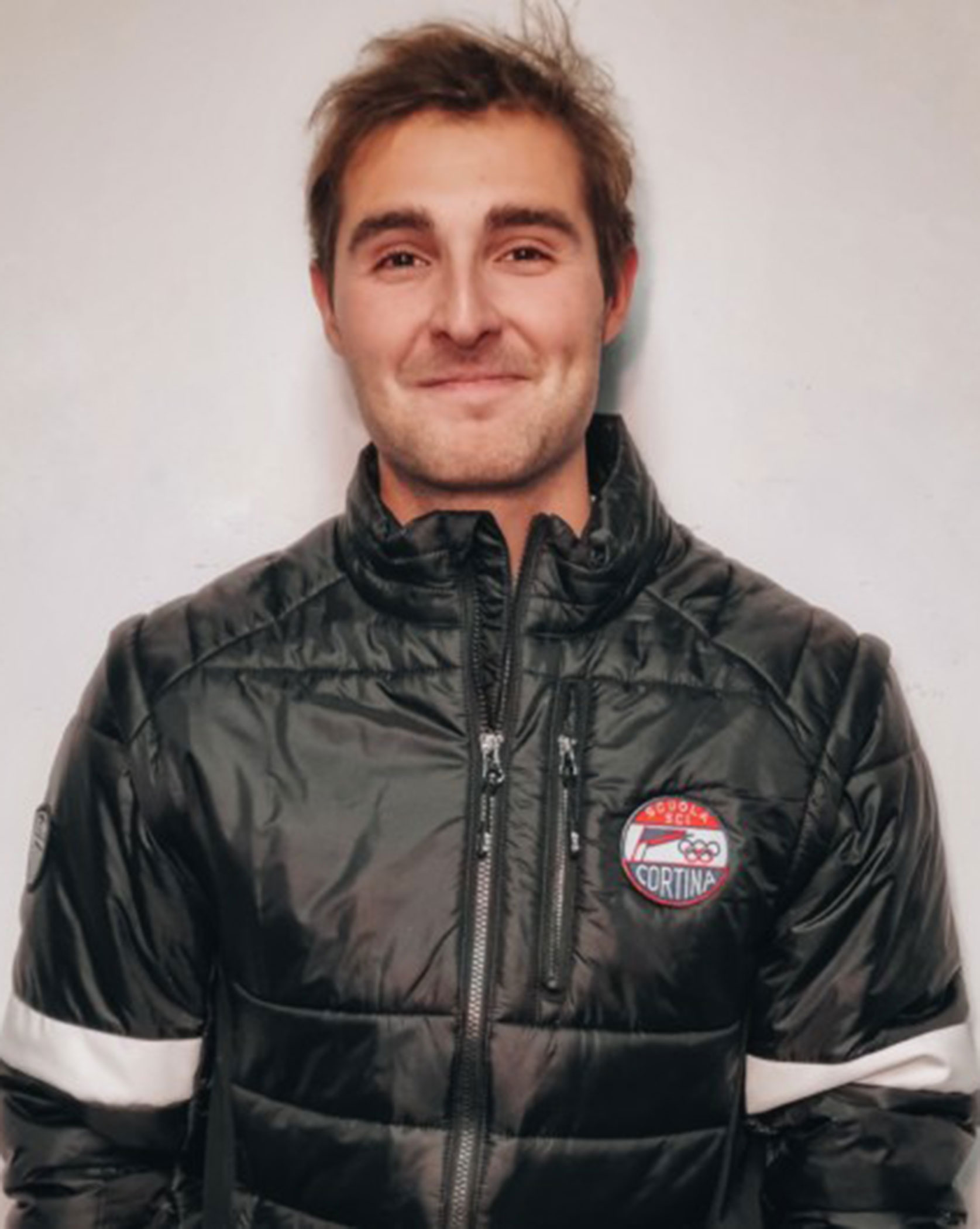 Smiling man in a black puffer jacket with a Cortina patch stands against a light-colored background. - Scuola Sci Cortina