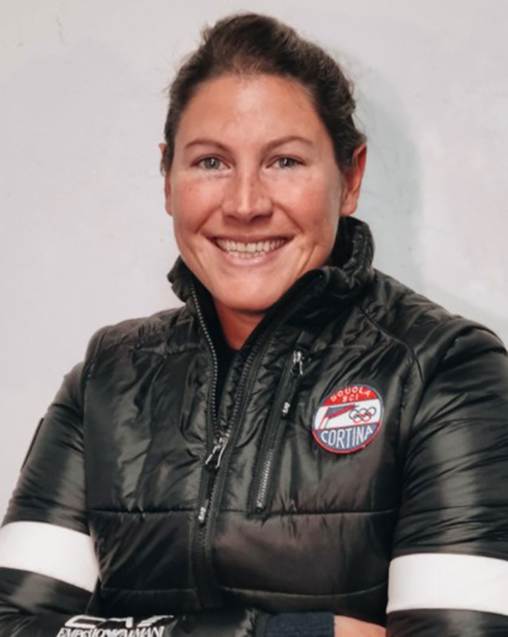 Woman in a black winter jacket with Cortina patch, smiling in front of a plain light background. - Scuola Sci Cortina