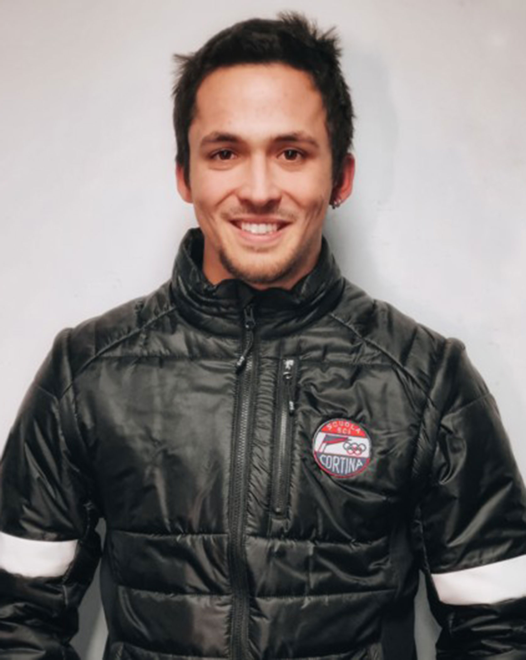 Smiling man in a black puffer jacket with a Cortina patch stands against a plain light background. - Scuola Sci Cortina