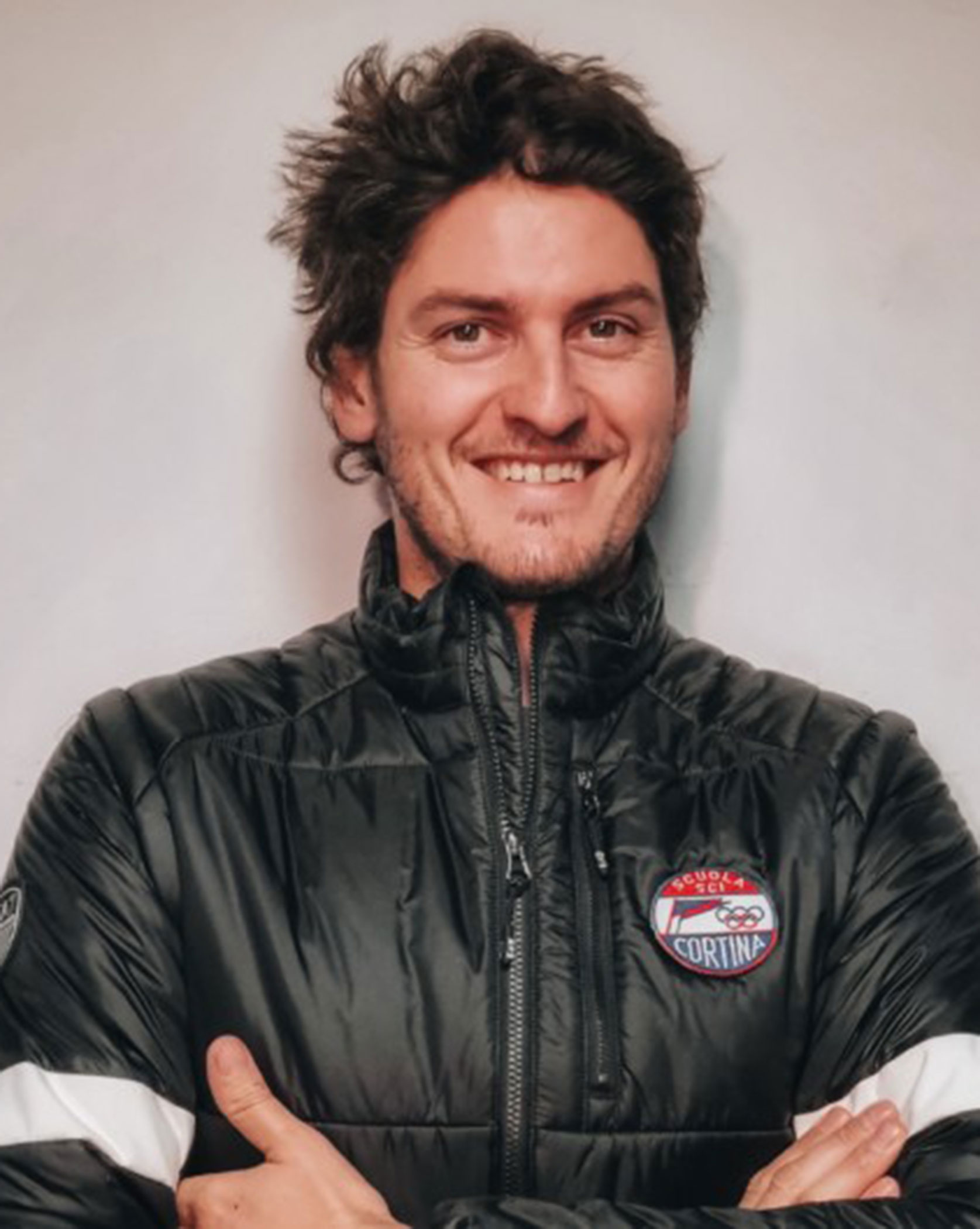 Man with wavy hair in a black jacket, smiling with arms crossed, standing against a plain background. - Scuola Sci Cortina
