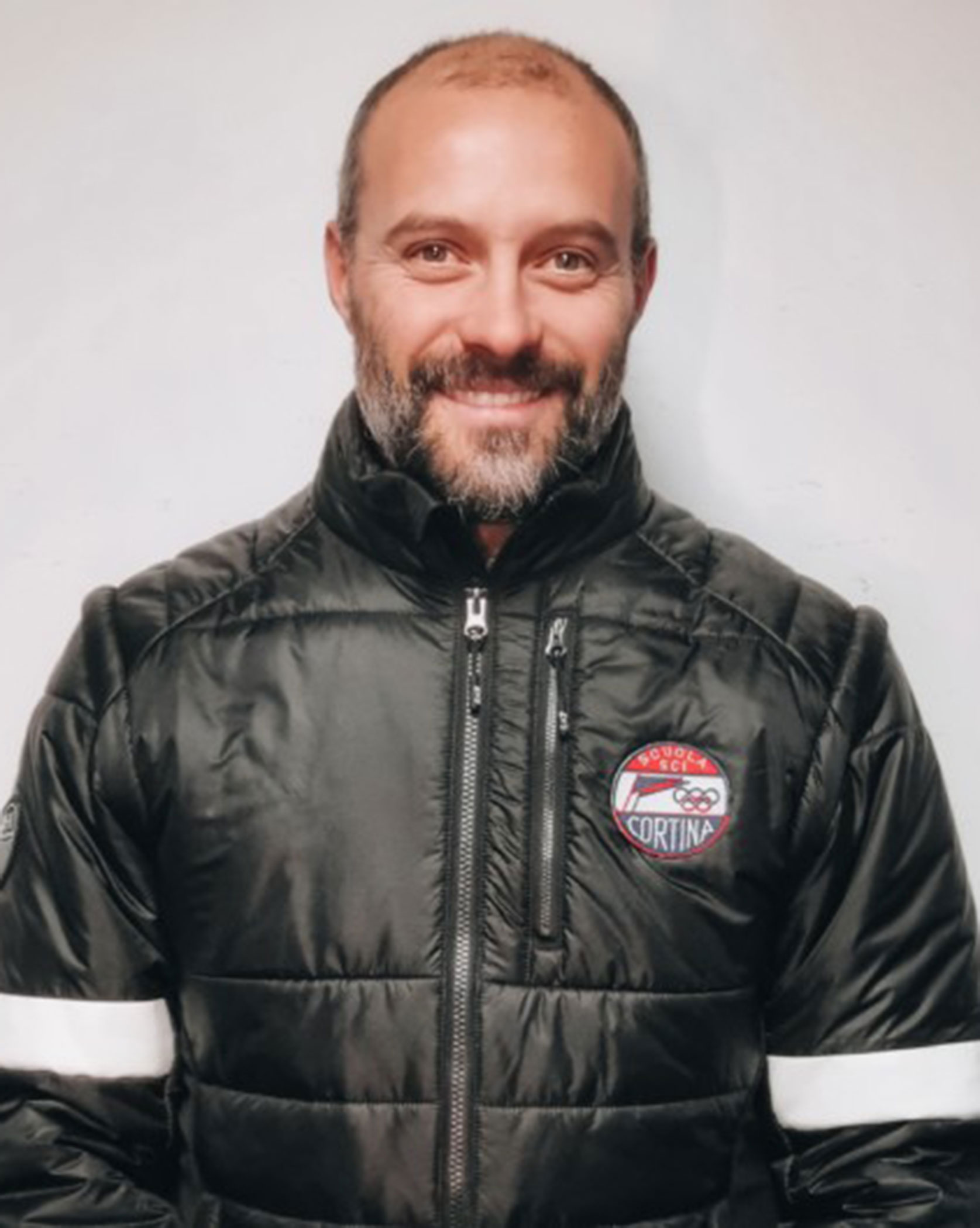Man with short hair and beard wearing a black jacket with a Cortina patch, smiling at the camera. - Scuola Sci Cortina