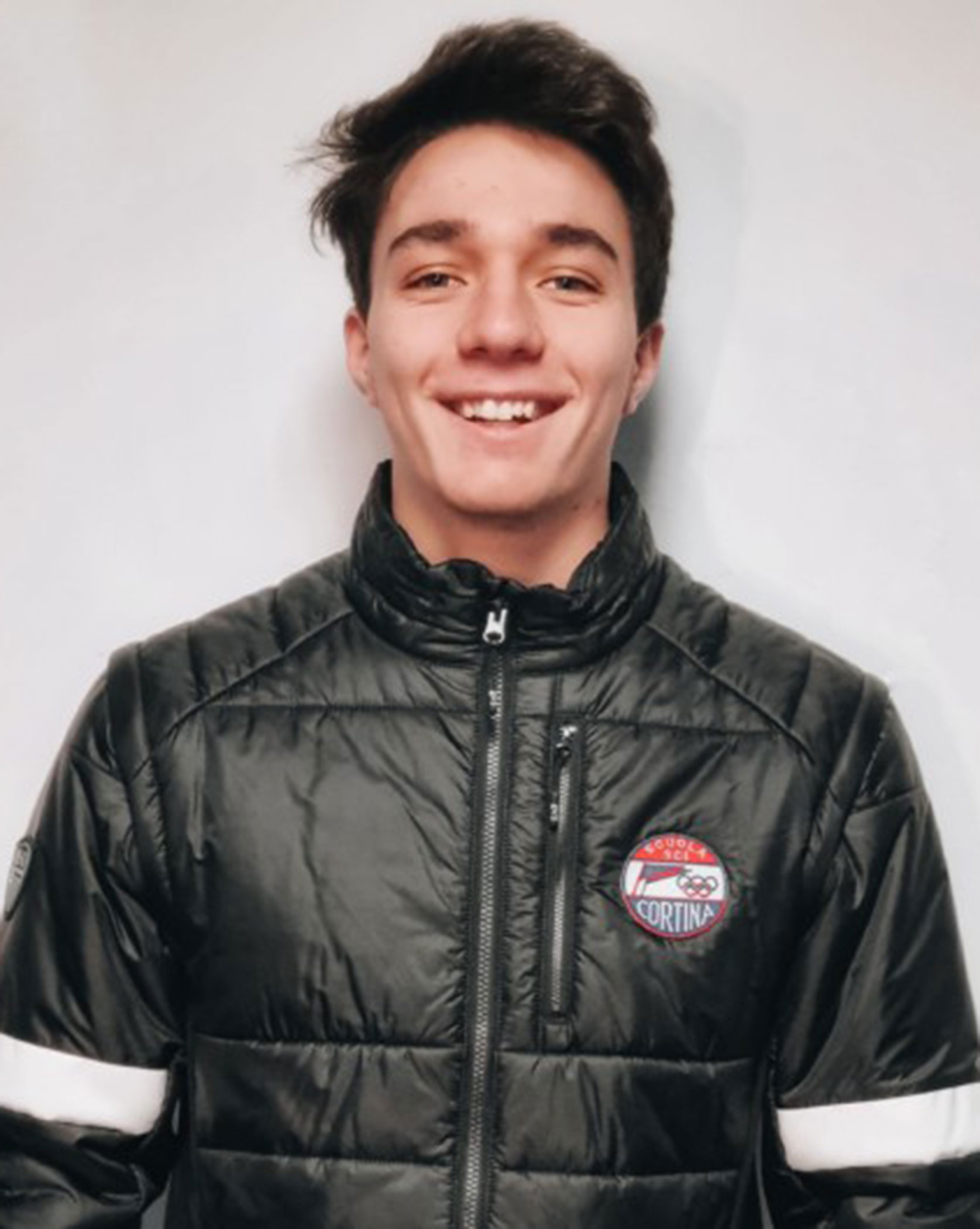 Young man in a black puffer jacket with a circular Cortina patch, smiling at the camera. - Scuola Sci Cortina