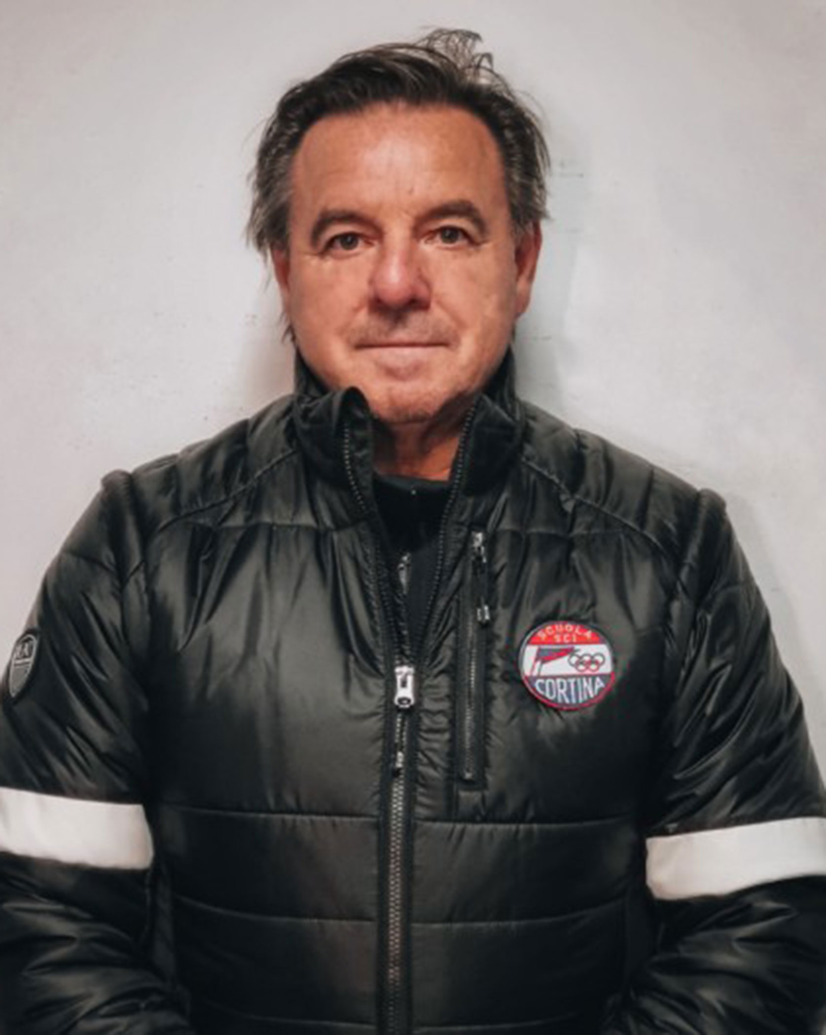 Man in a black puffer jacket with a Cortina patch, standing against a plain light background, looking forward. - Scuola Sci Cortina