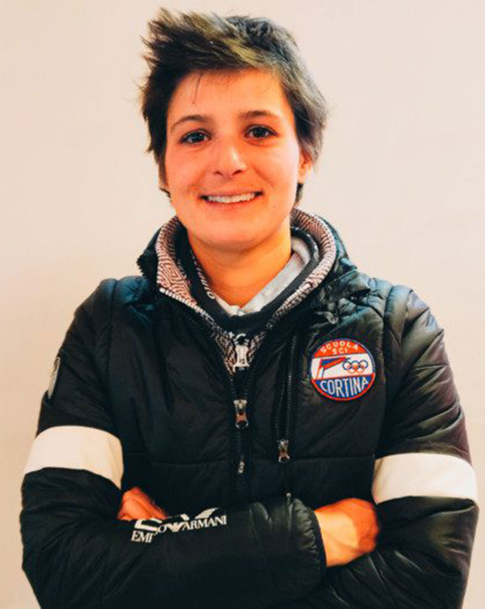 Person with short hair smiling, arms crossed, wearing a black jacket with a Cortina badge on it. - Scuola Sci Cortina